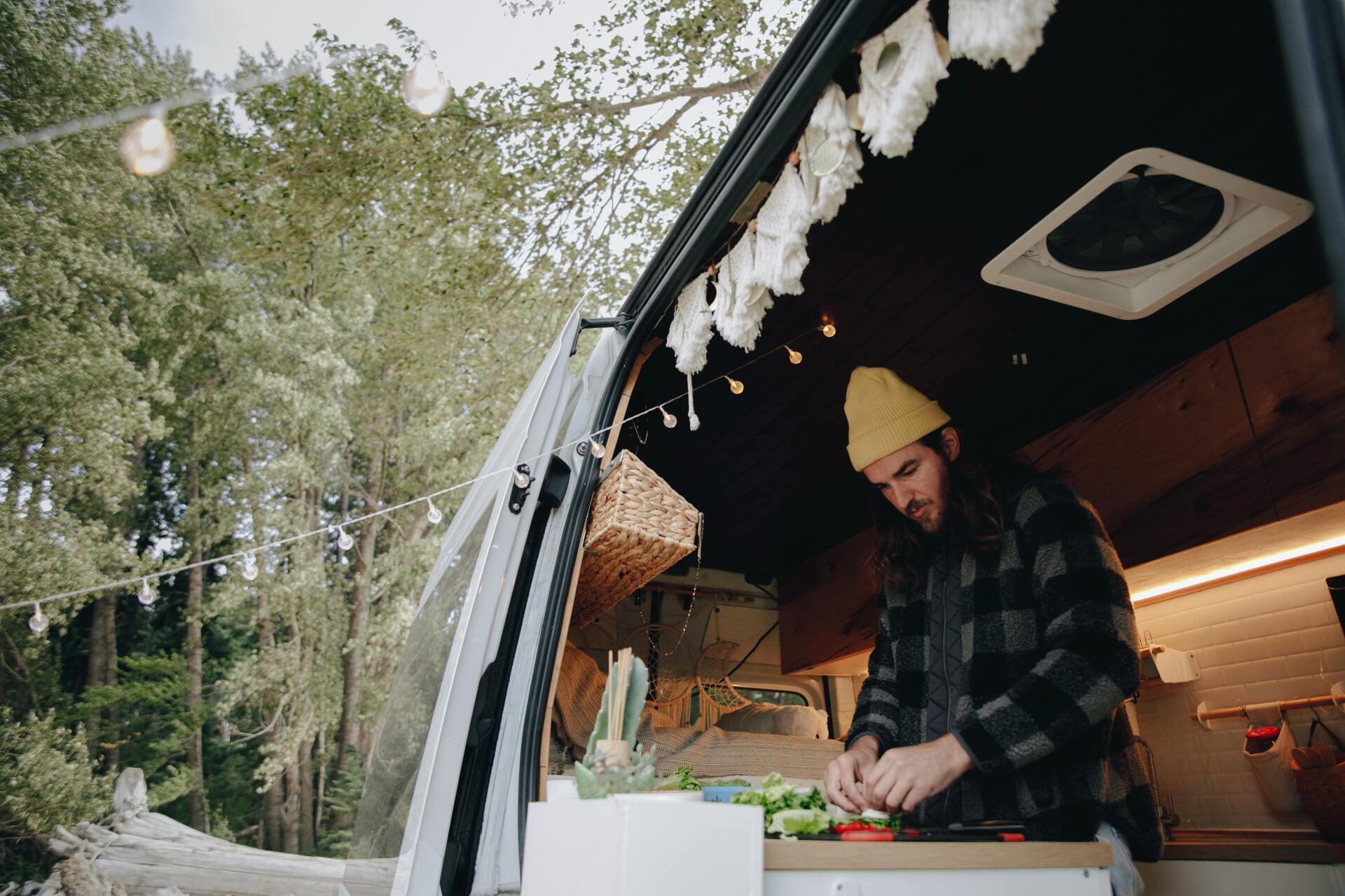 Man cooking inside a camper van, outdoors; wearing a yellow beanie and plaid shirt.