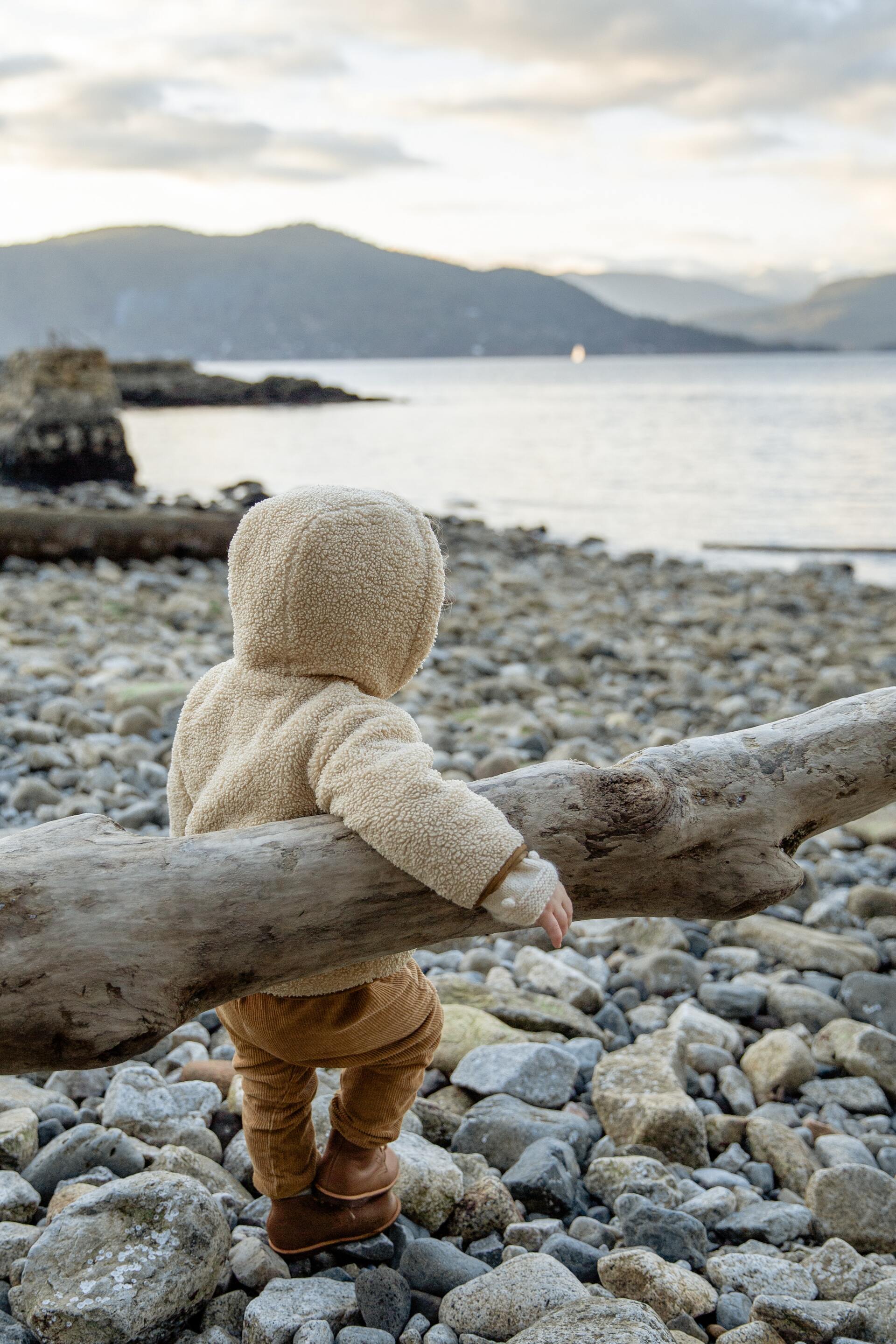 Child in a hooded jacket looking out at a calm sea from a rocky beach, leaning on a log.