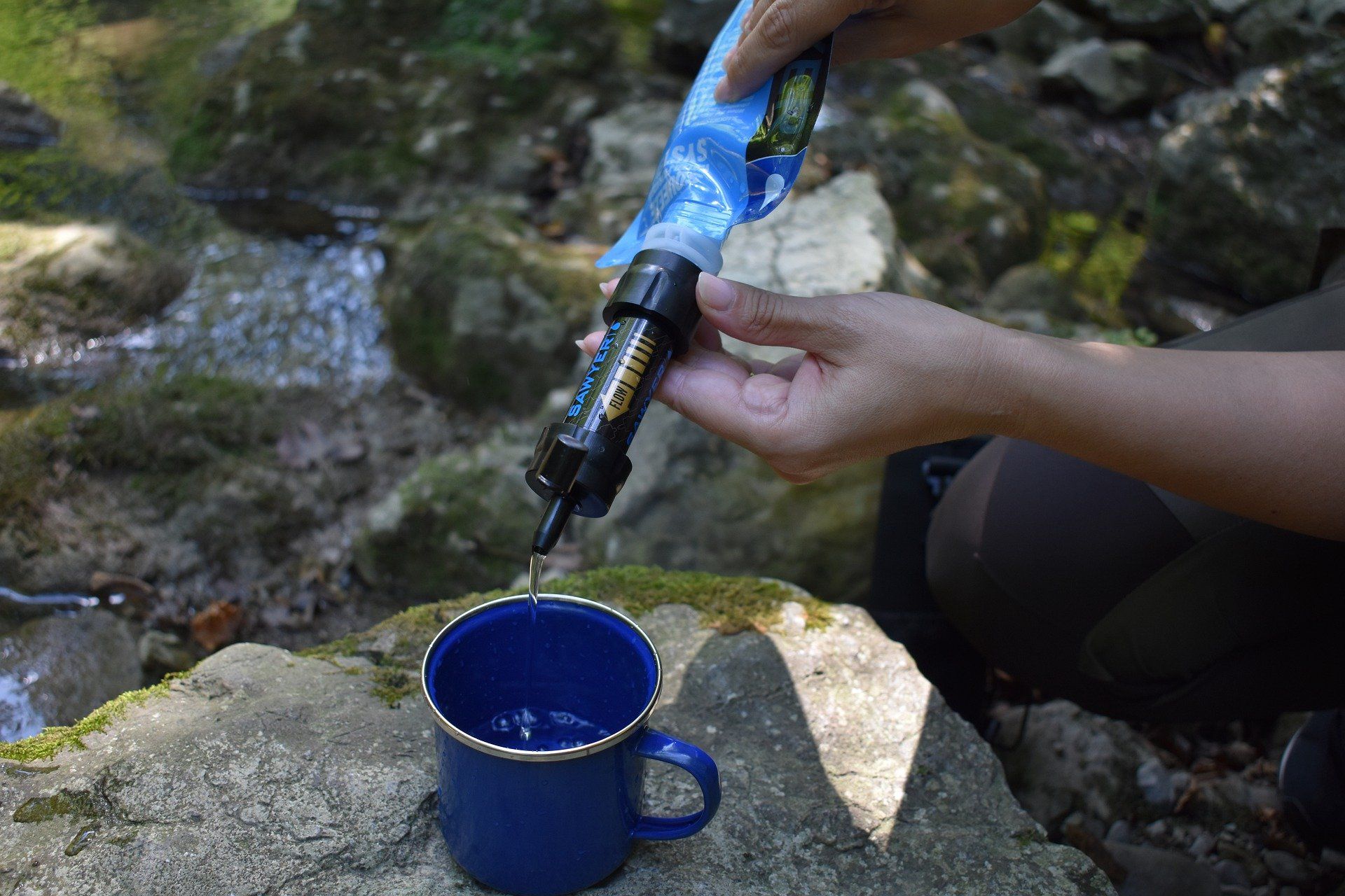 Person filtering water from a stream into a blue mug using a portable water filter.