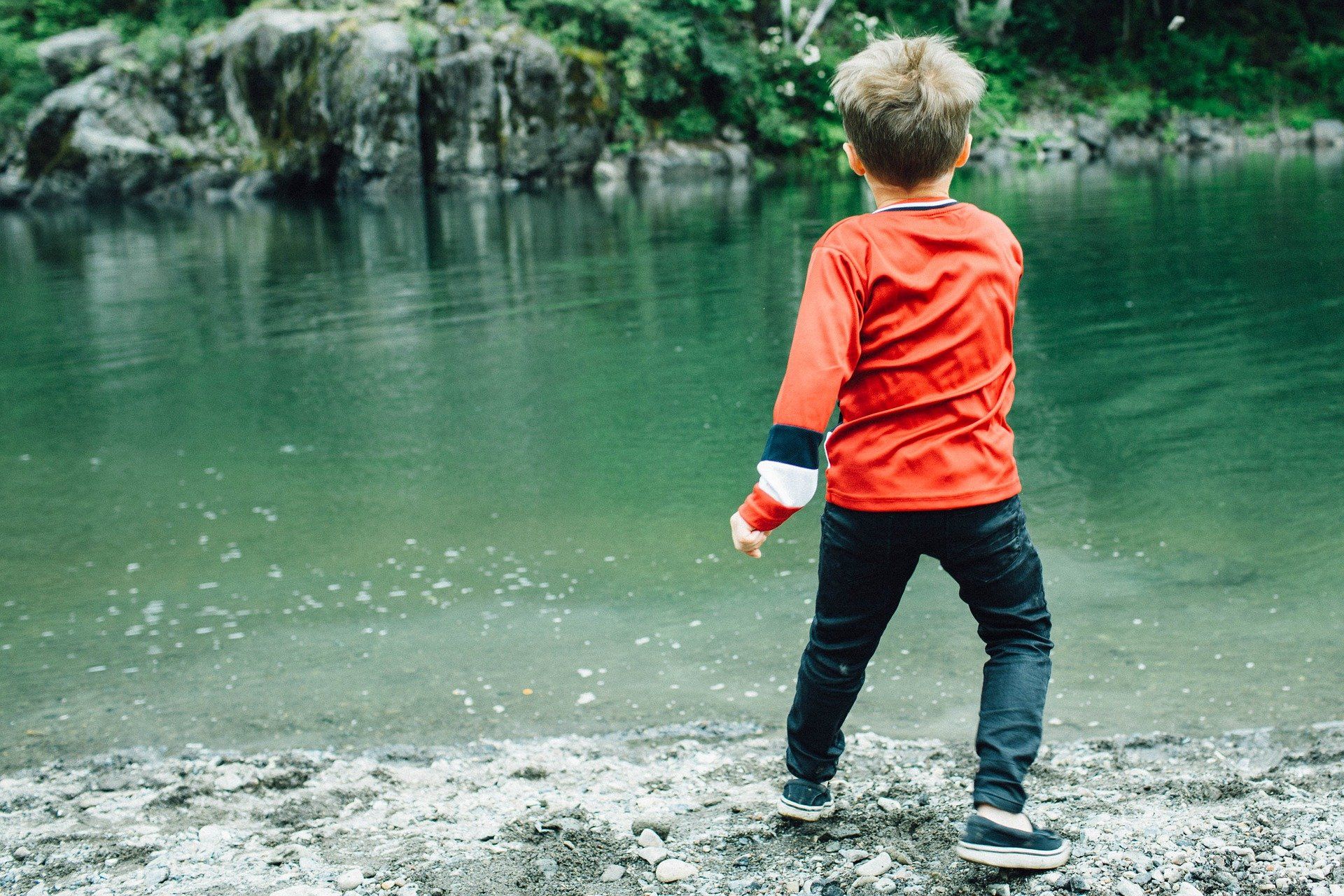Boy in red shirt and dark jeans throws stones into a lake.