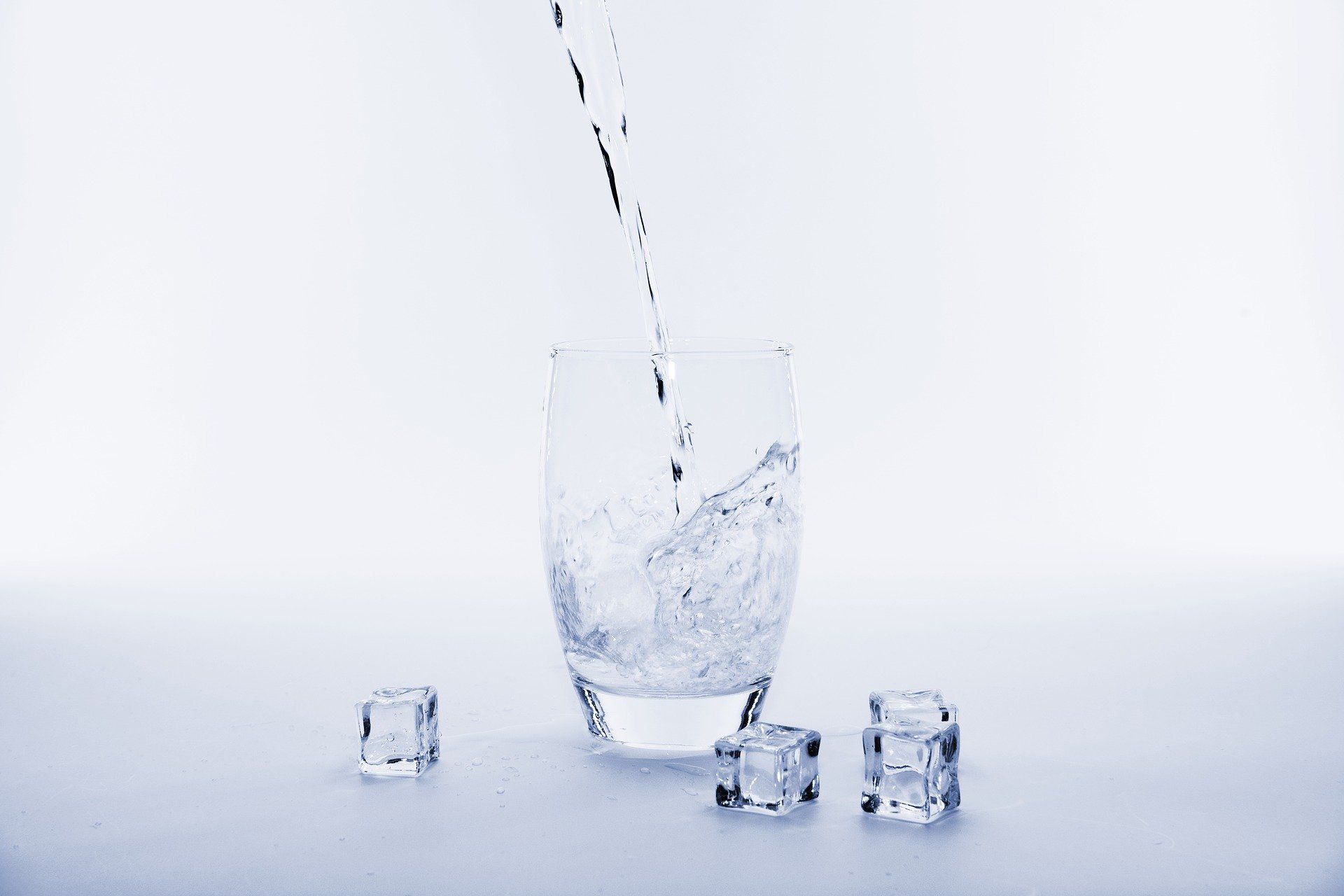 Water being poured into a glass, splashing, with ice cubes on a white surface.