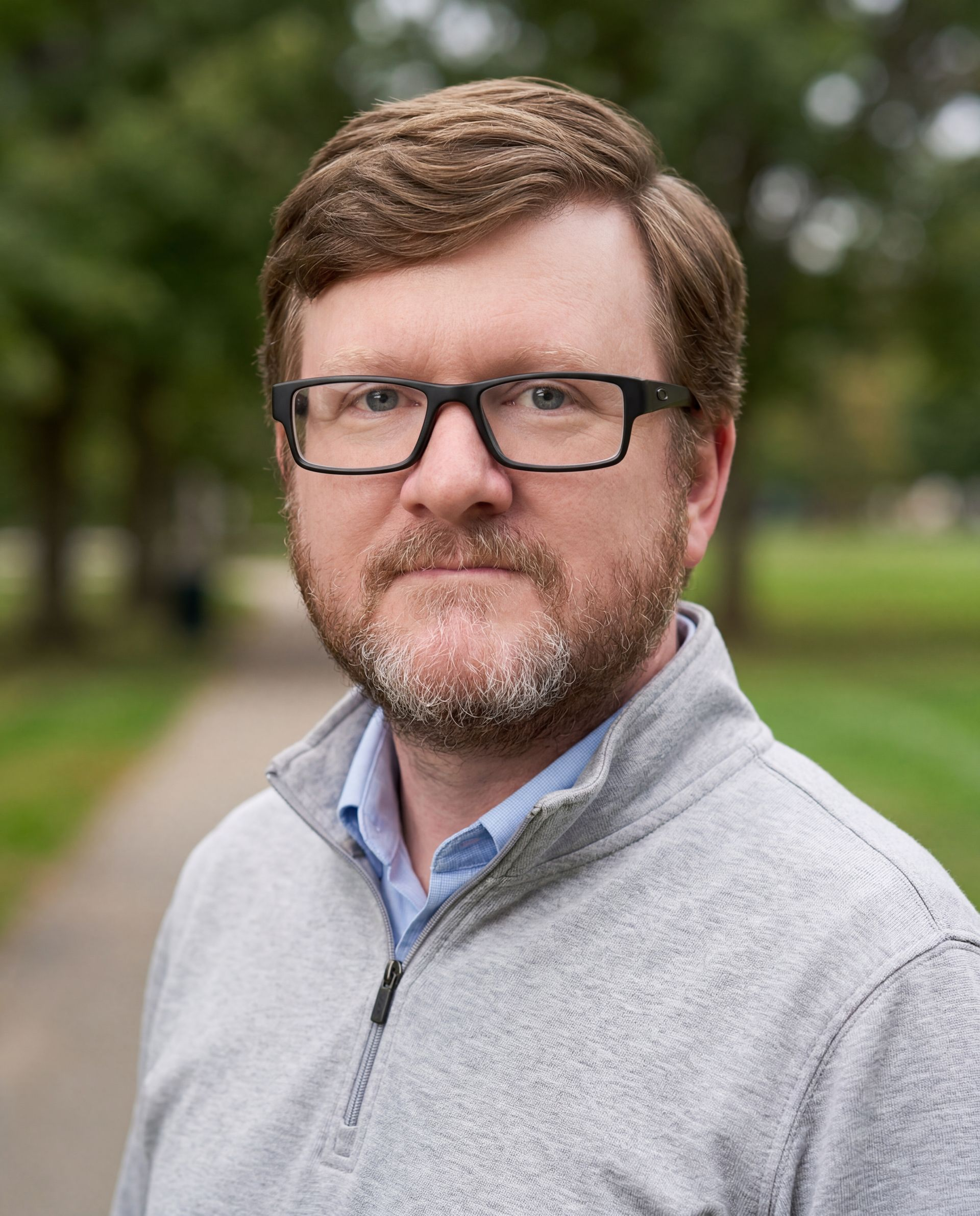 A person with reddish-brown hair and glasses, wearing a grey quarter-zip sweater, stands outdoors in a park setting.