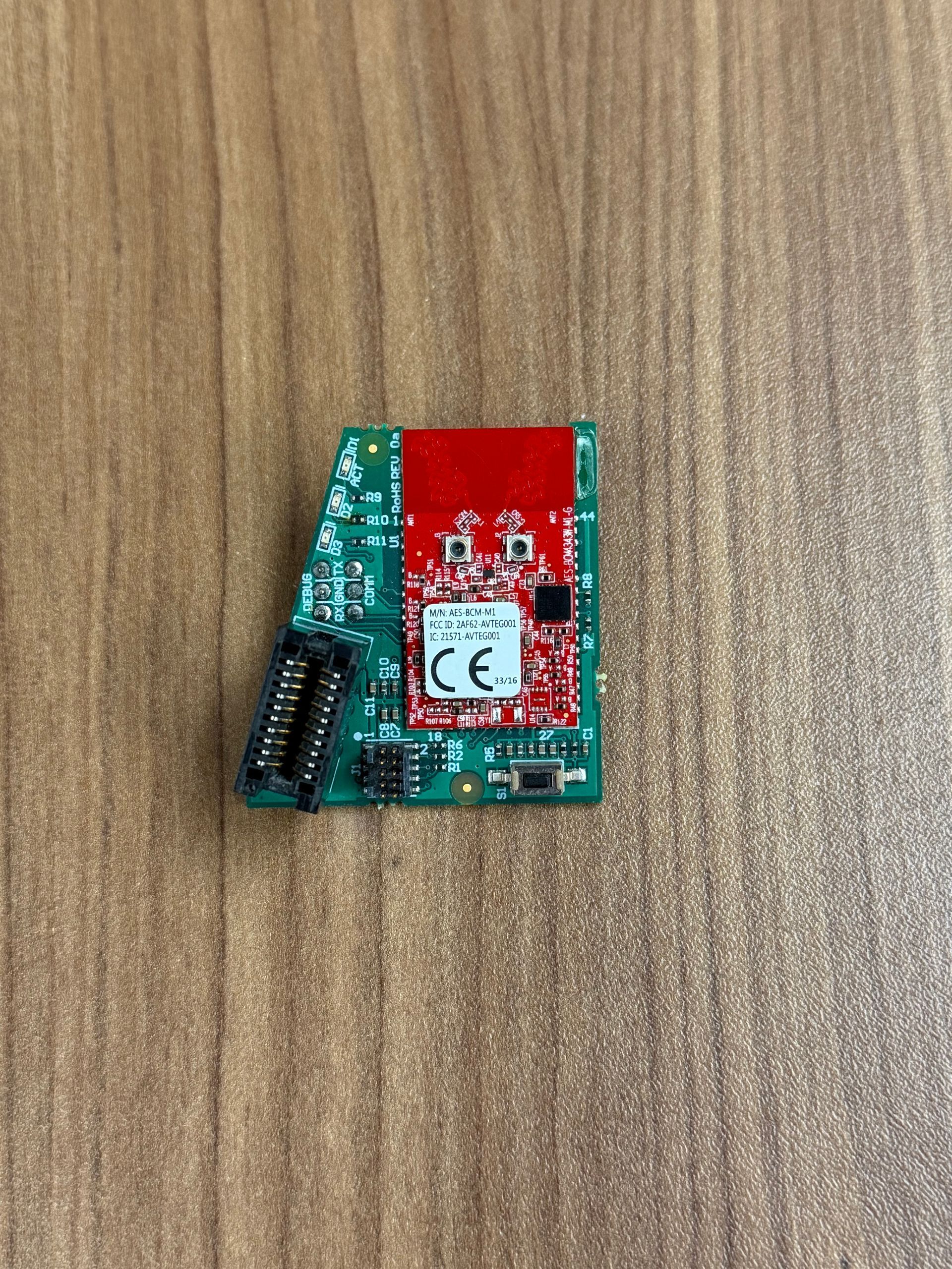 A small, rectangular green circuit board with a red wireless module component sitting on a wooden surface.