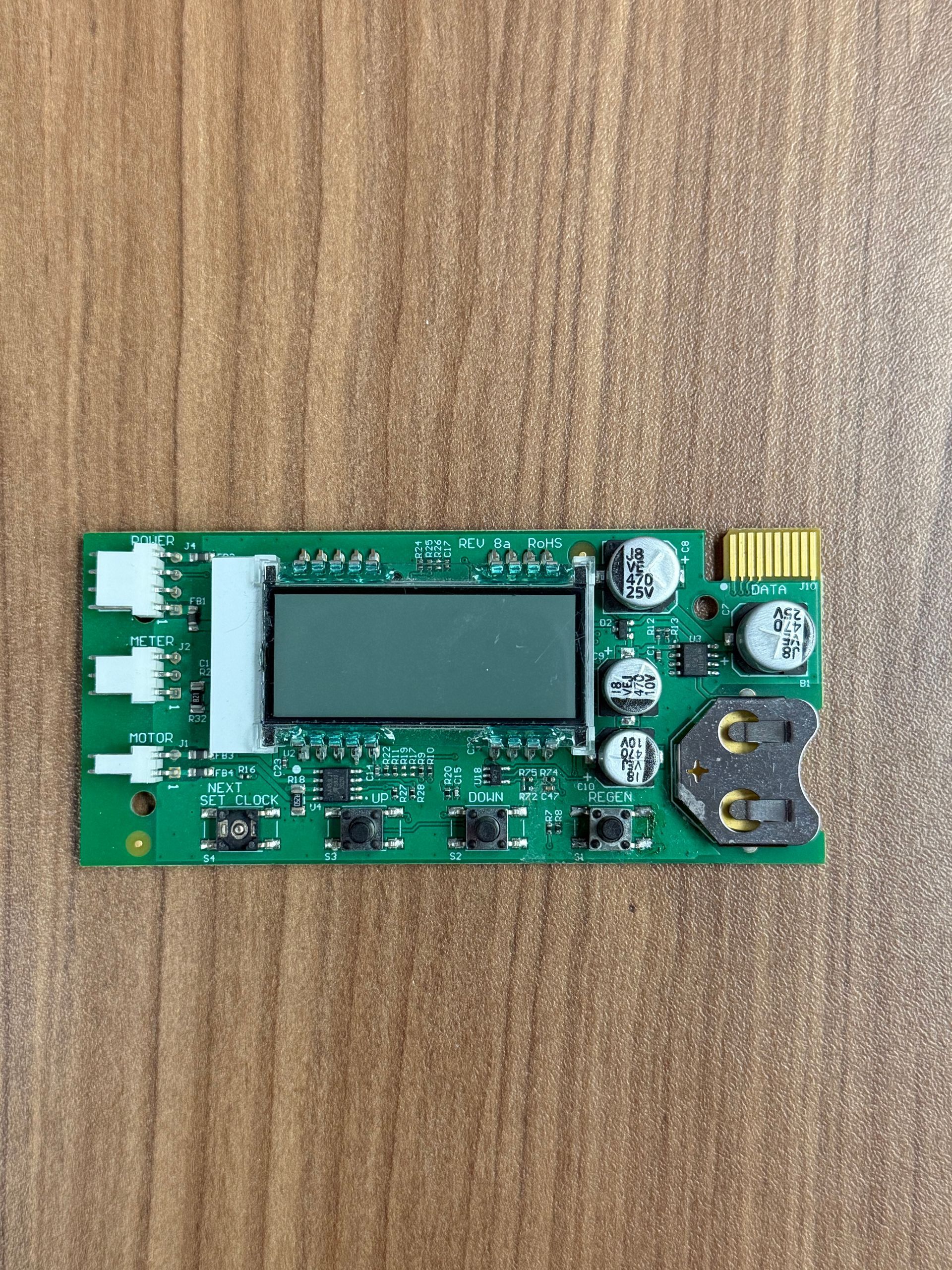 A green circuit board featuring a rectangular LCD screen, small buttons, and a coin cell battery holder on a wood surface.