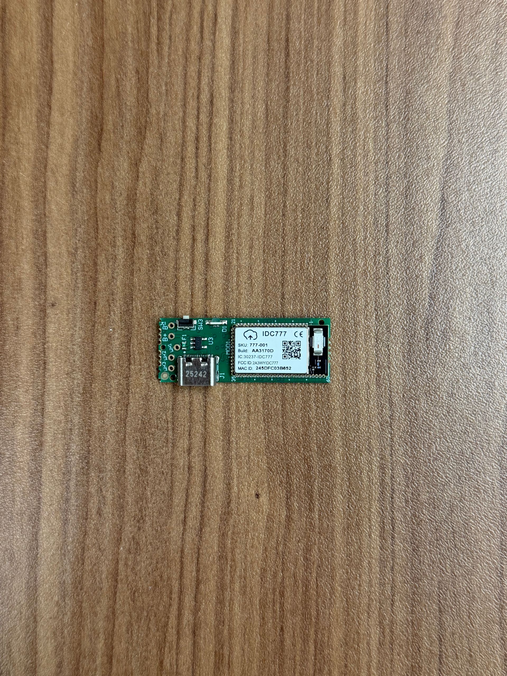 A small green circuit board with a USB-C port and a rectangular wireless module, centered on a wood-grain surface.