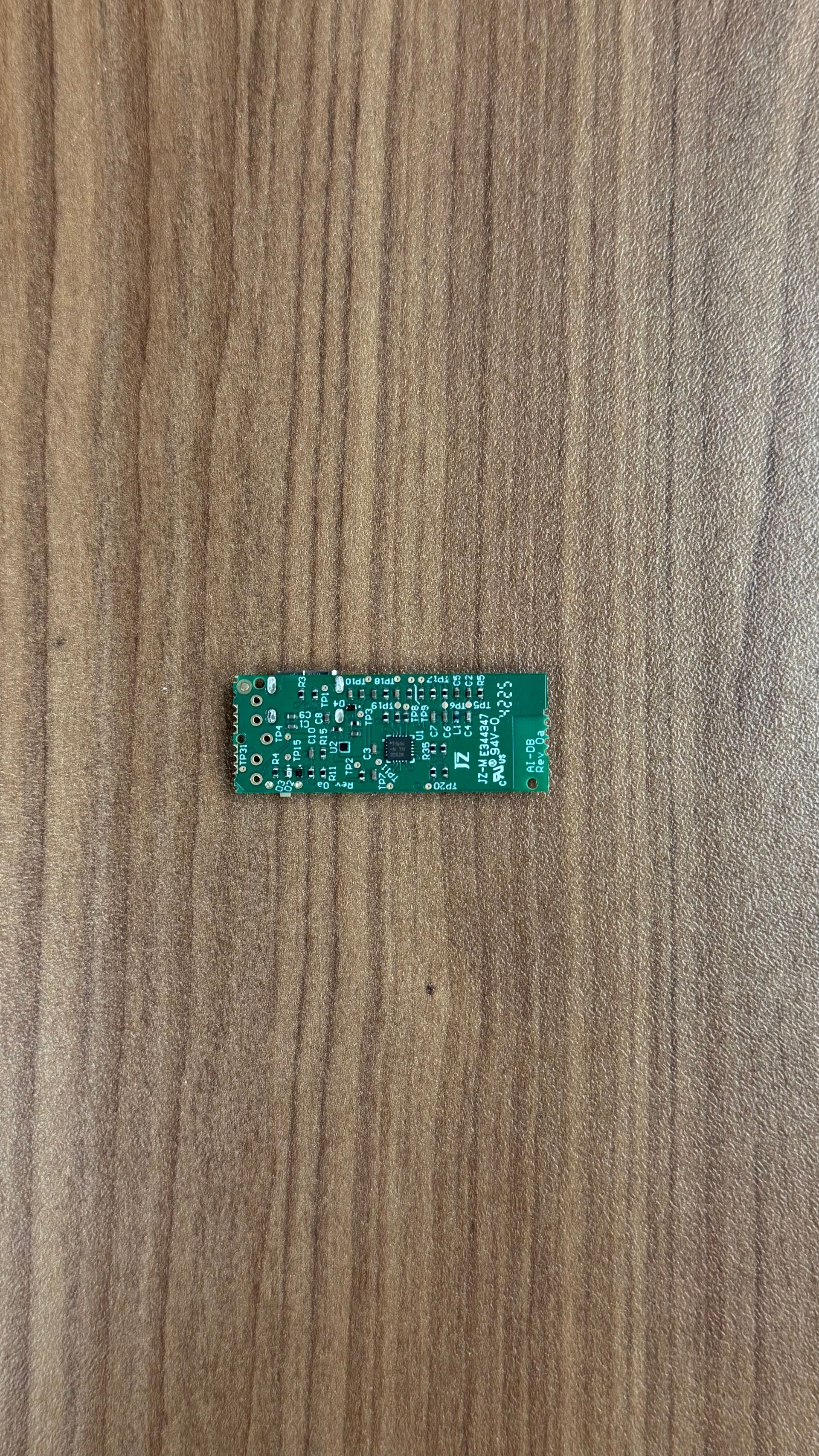 A small, rectangular green circuit board with tiny silver electrical components lies on a textured wood-grain surface.