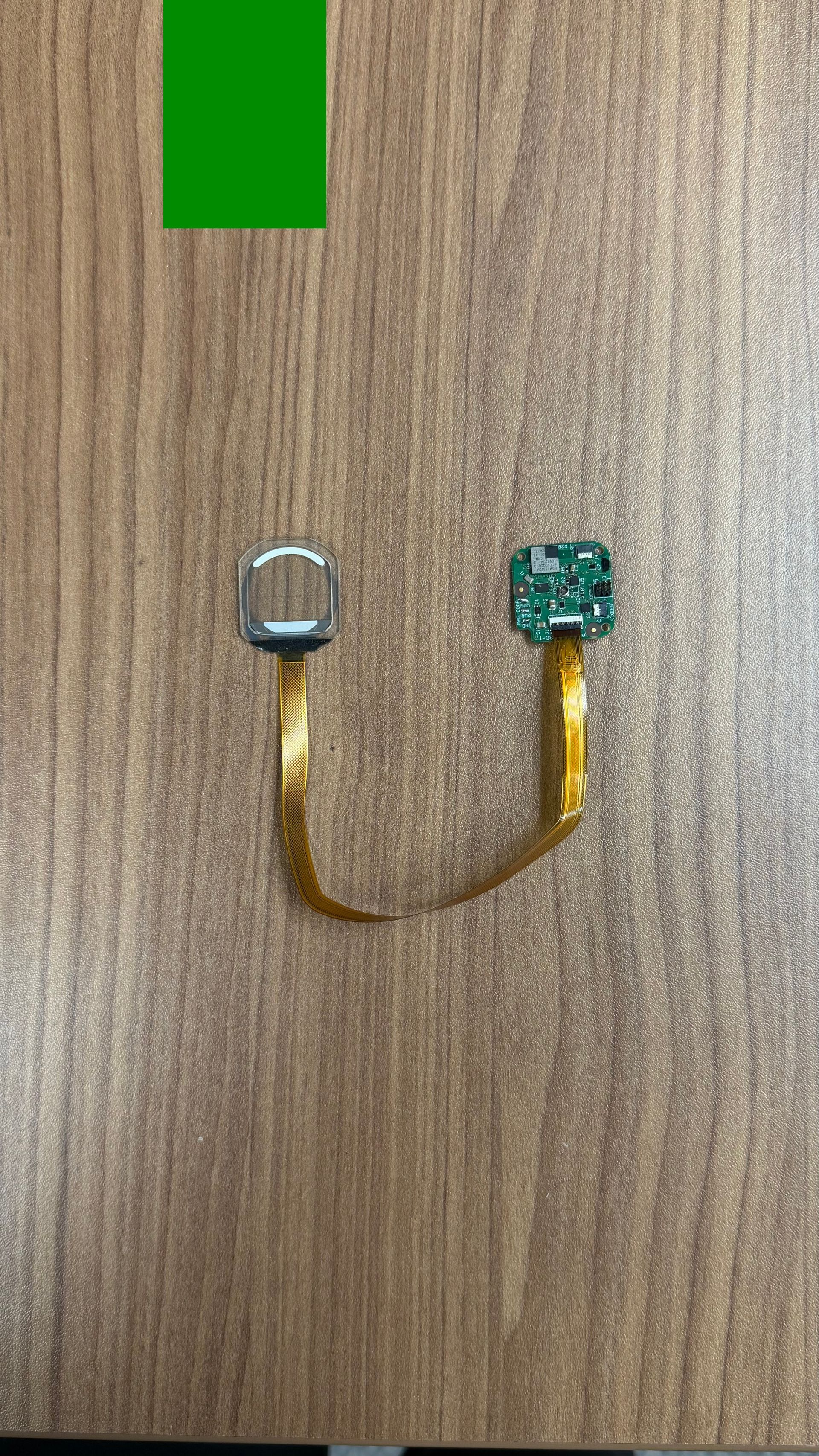 Two small square components—one silver, one green—connected by a thin, flexible gold ribbon cable on a wood grain surface.