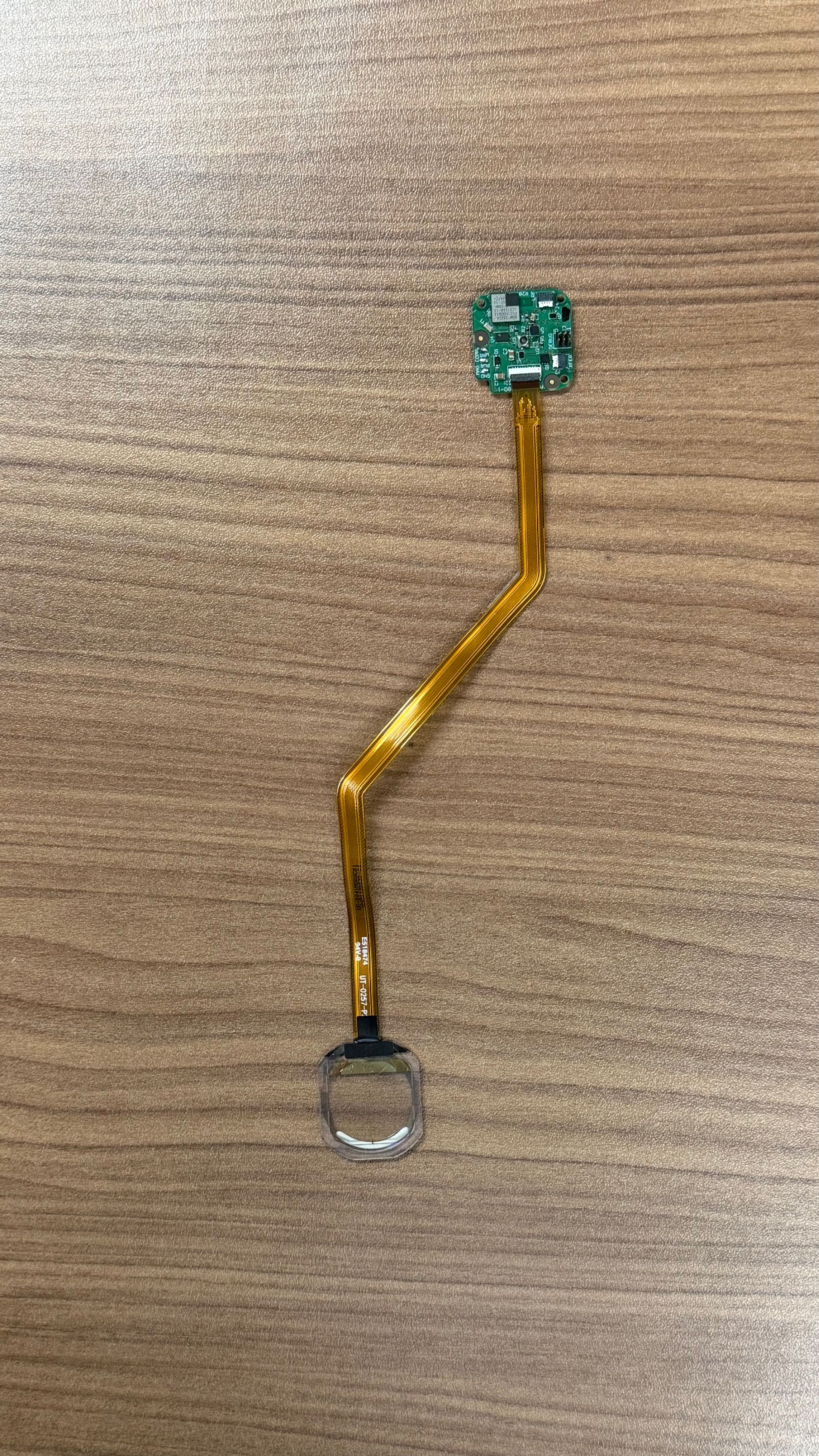 A small electronic fingerprint sensor module with a thin, flexible gold-colored ribbon cable, lying on a wooden surface.