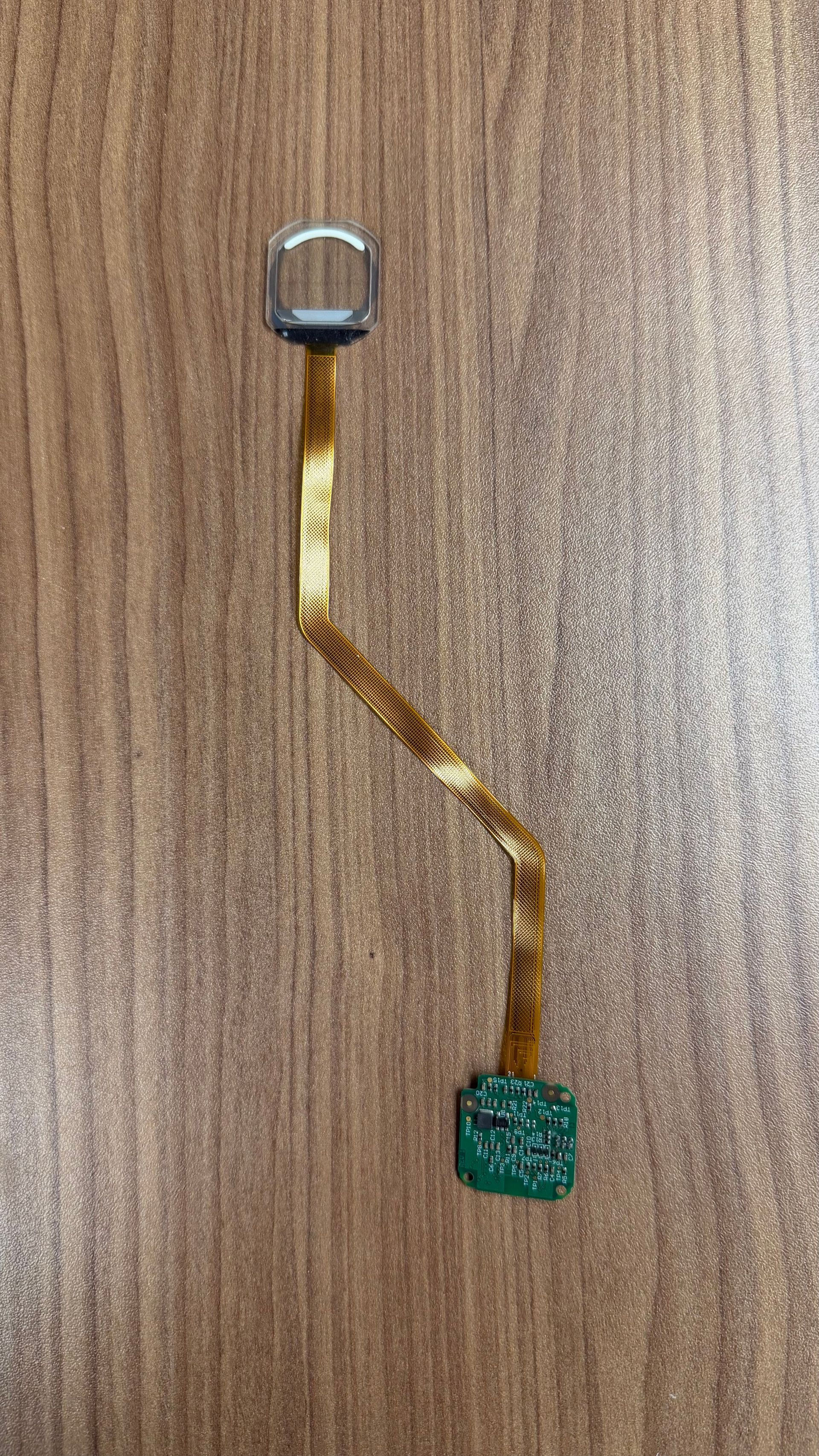 A small, green electronic sensor module attached to a long, thin, flexible golden ribbon cable on a wood-grain surface.