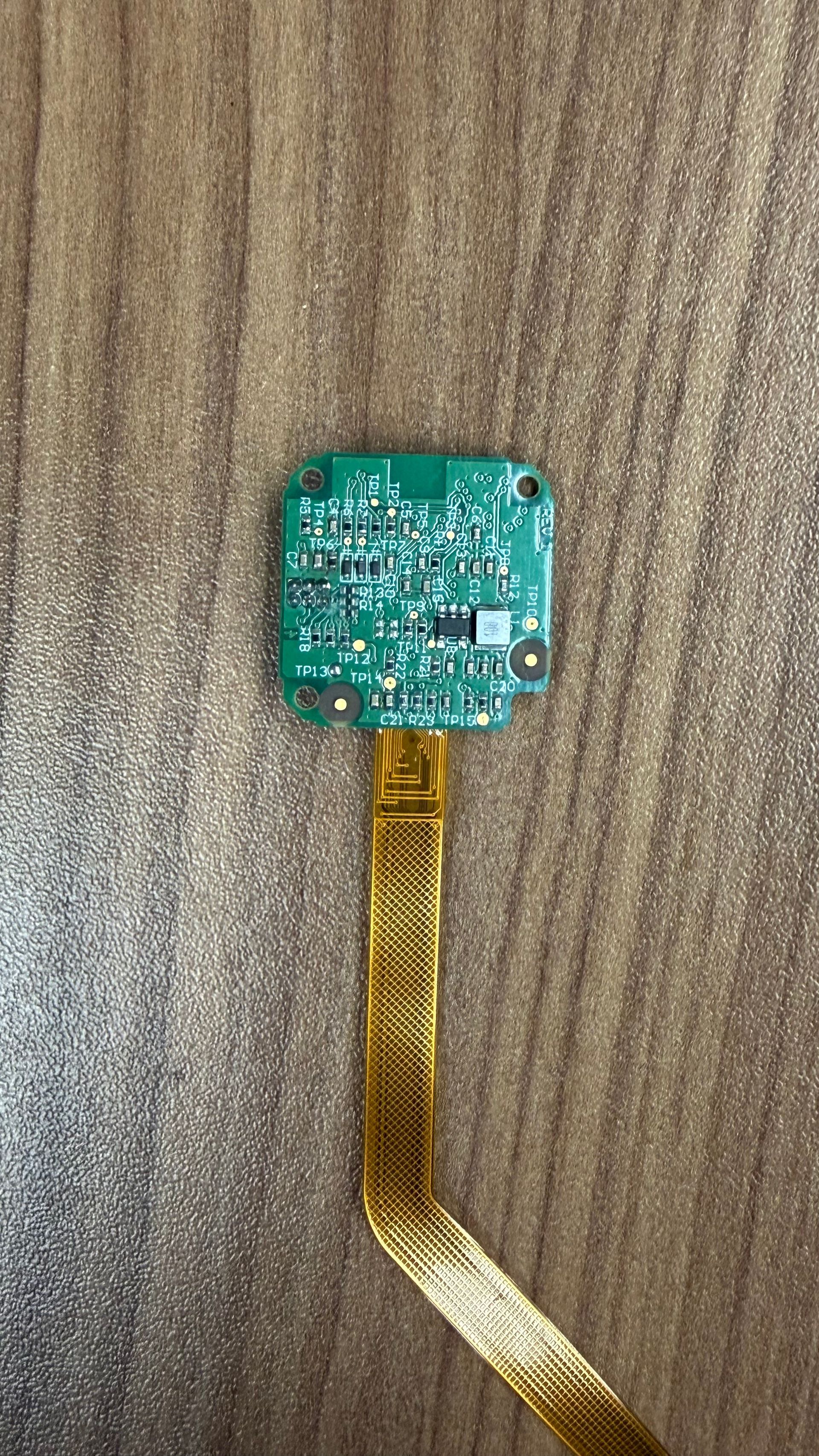 A small, square green circuit board attached to a flexible, gold-colored ribbon cable, resting on a wood-grain surface.