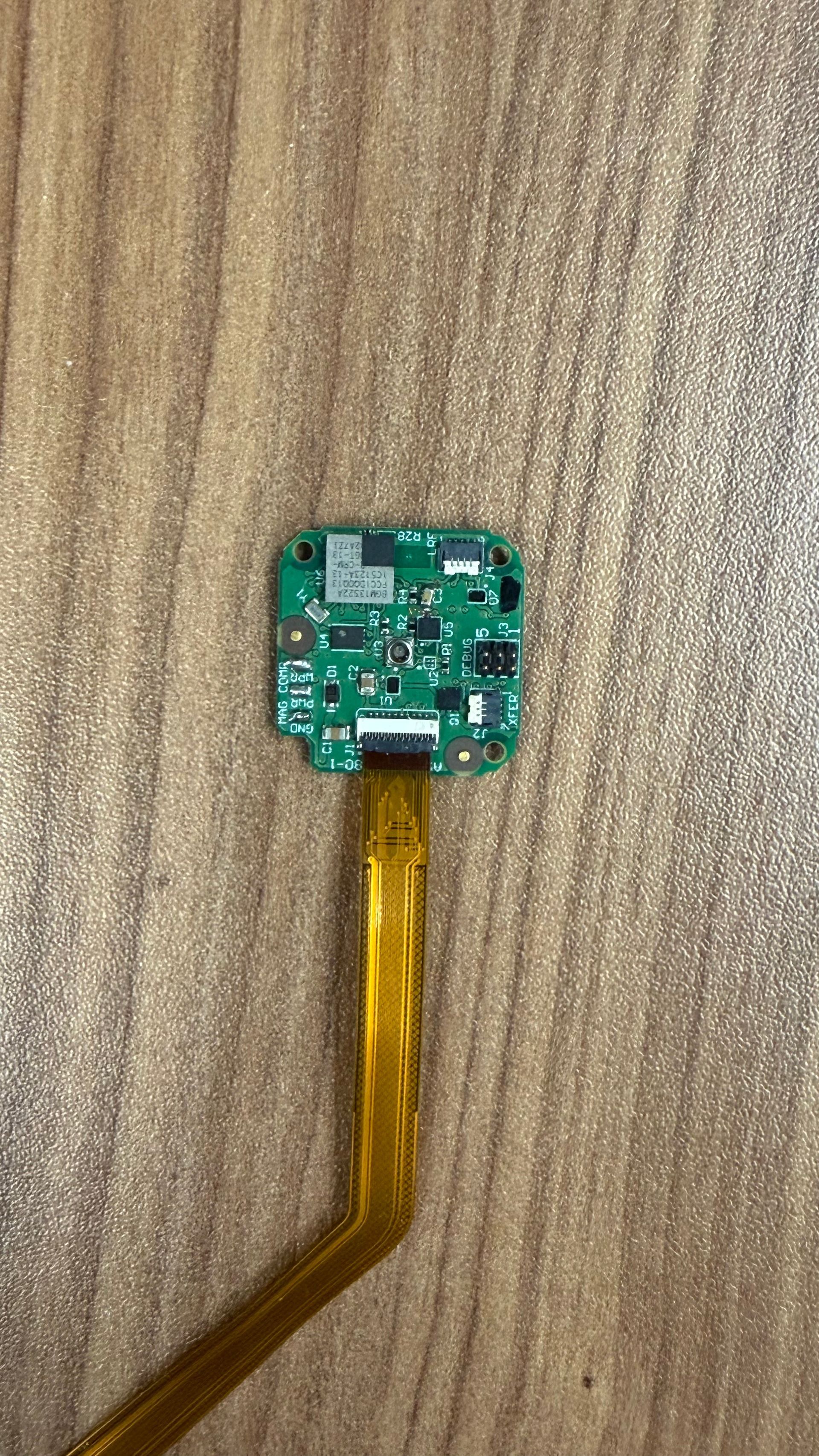 A small, green square circuit board attached to a long, flexible amber ribbon cable against a wooden surface.