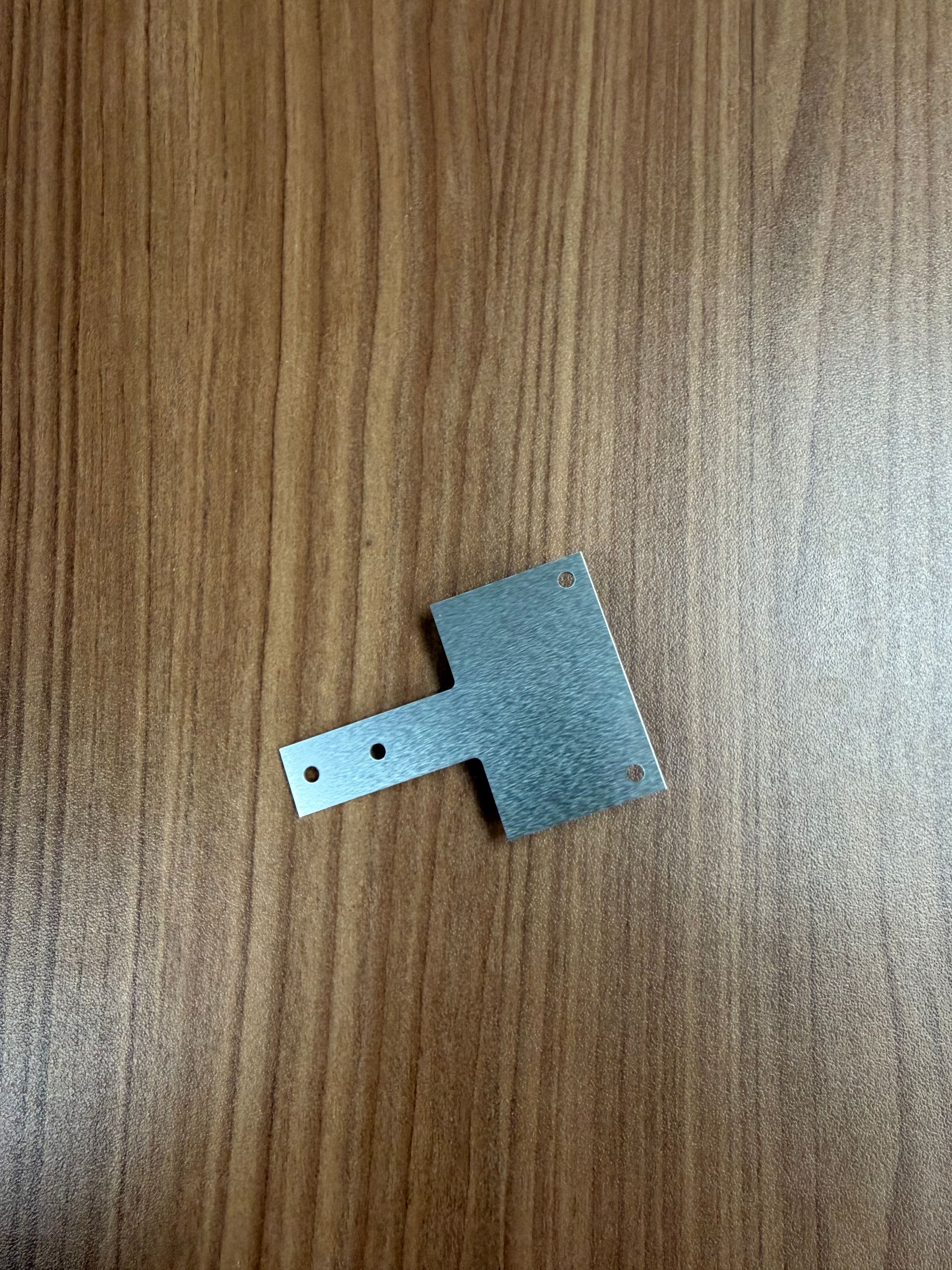A small, T-shaped metal bracket with two screw holes on its narrow arm, resting on a brown wood-grain surface.