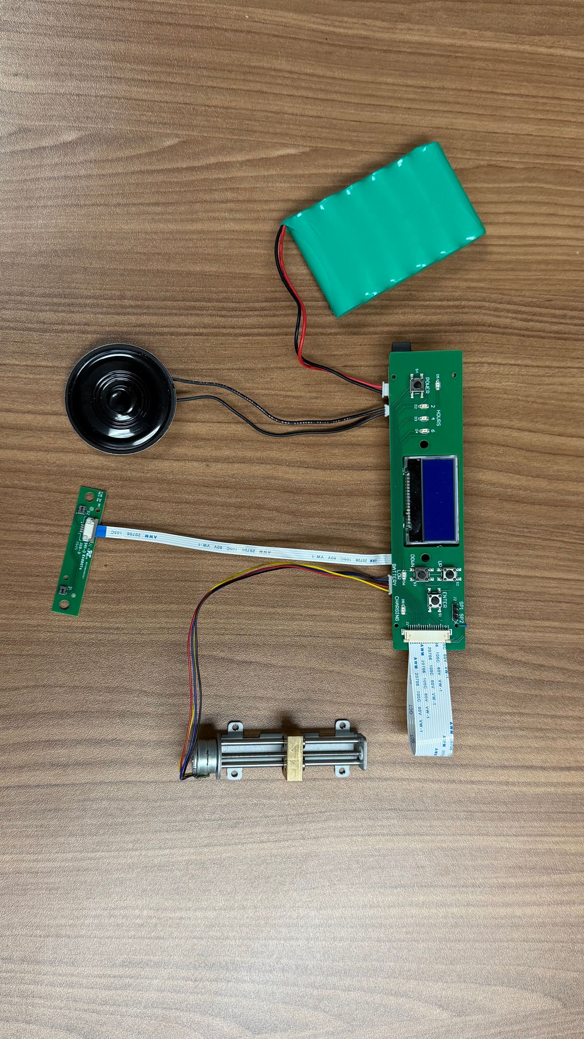 Disassembled electronic device with a green circuit board, speaker, battery pack, and a metal component on a wooden desk.