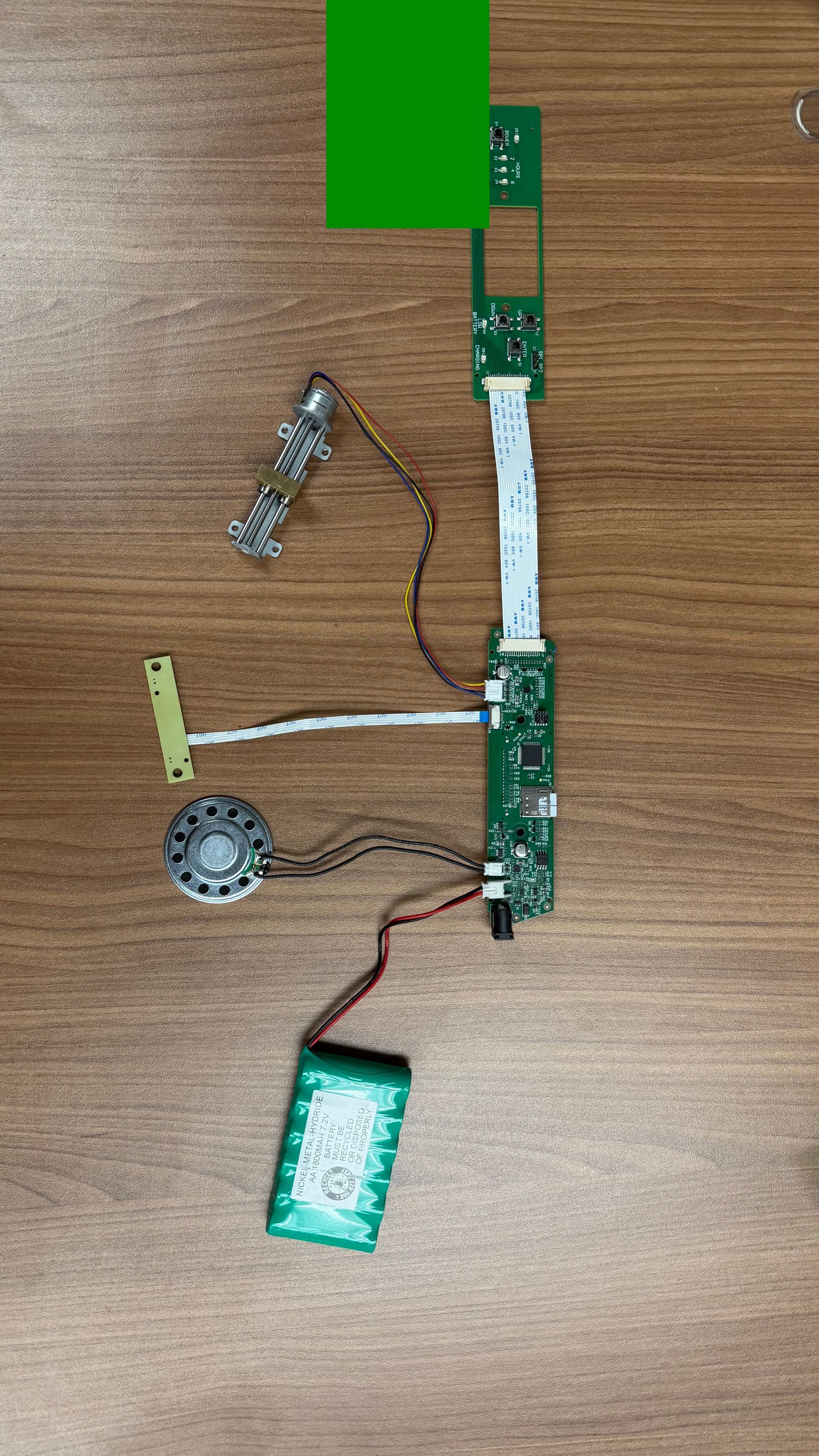 Disassembled electronics with a green circuit board, speaker, battery, and ribbon cable arranged on a wooden surface.
