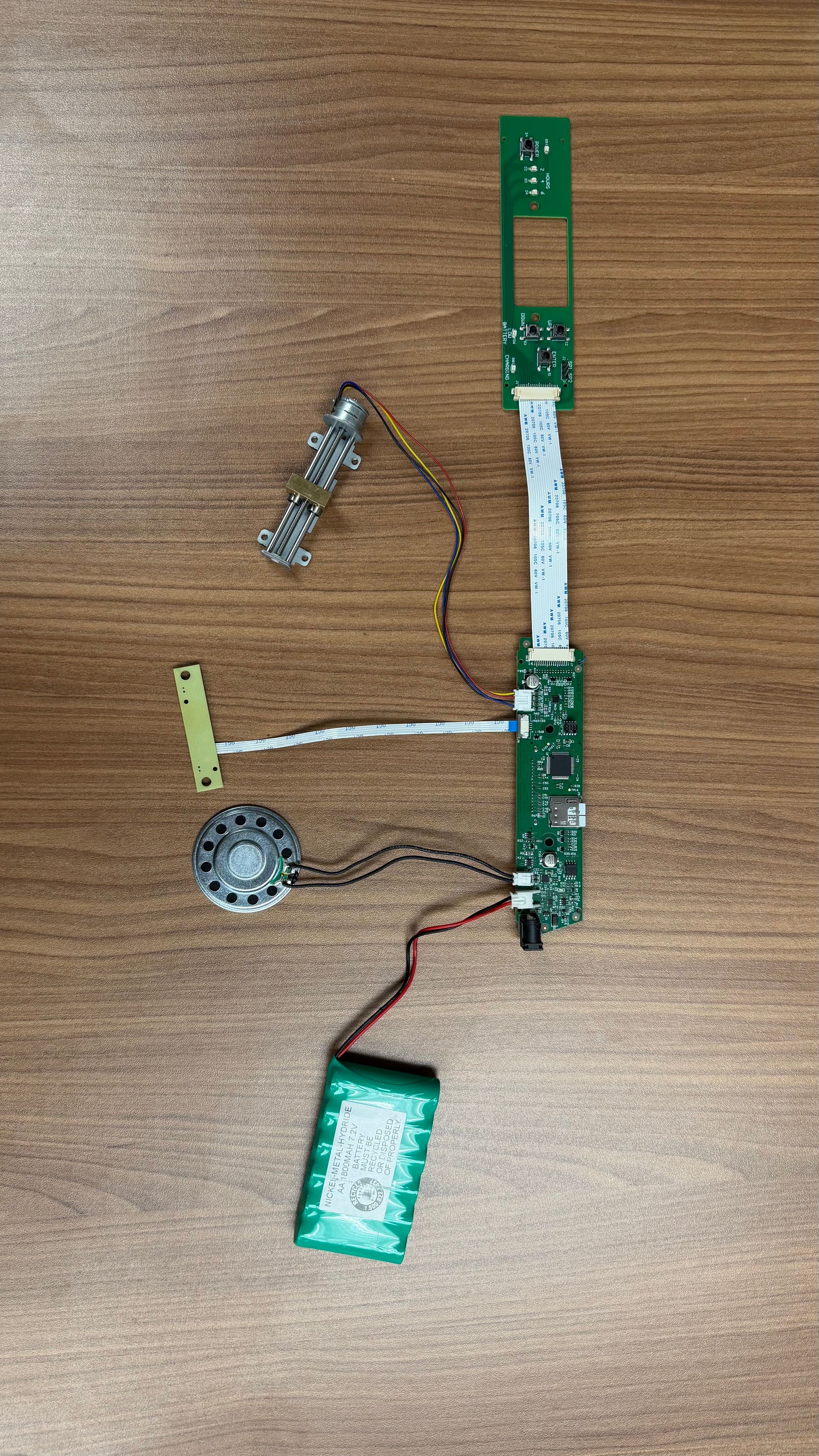 An assembled green circuit board with a small speaker, battery, and connecting wires on a brown wooden surface.