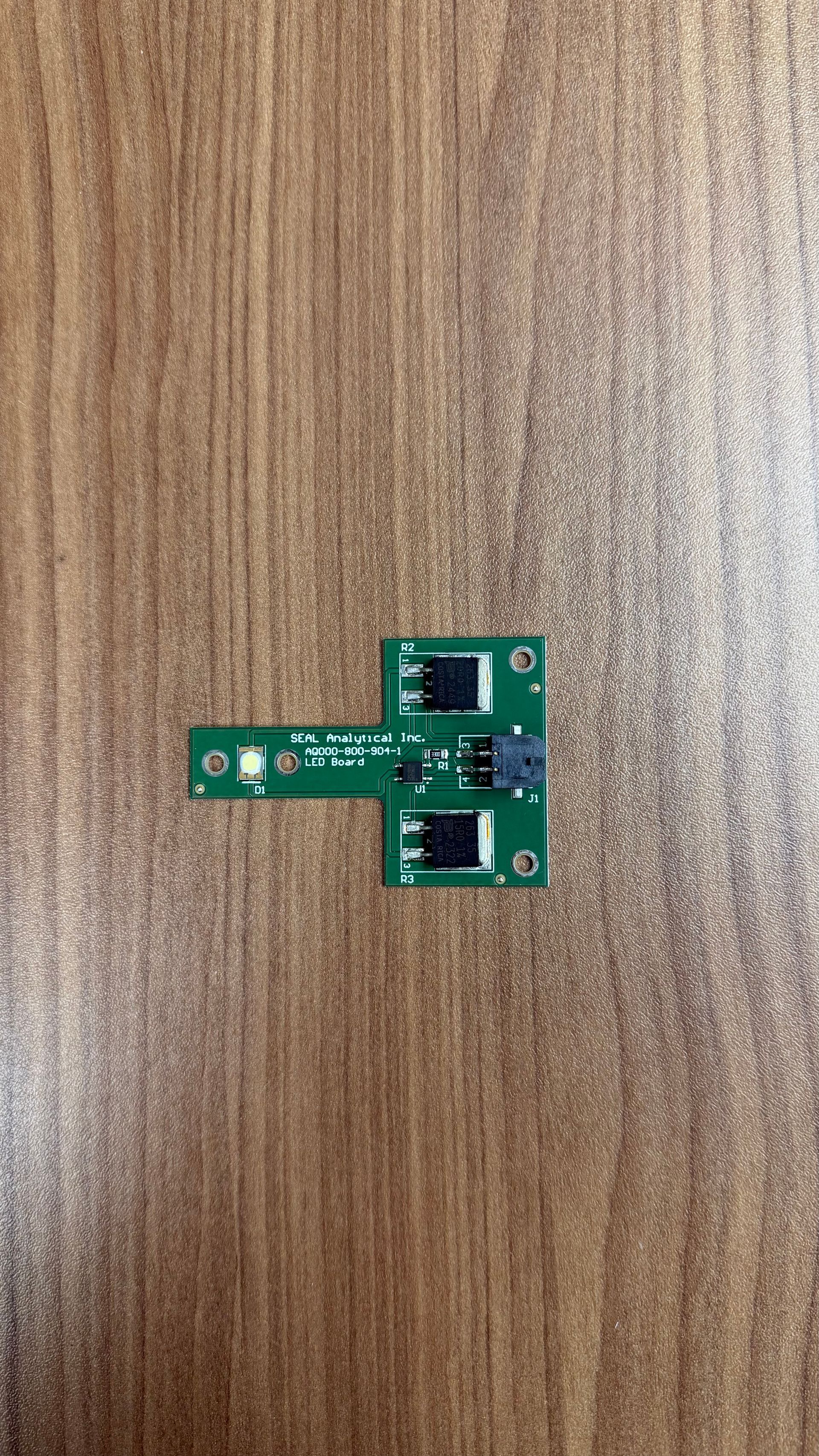 A small T-shaped green circuit board with two black sensors and a white connector on a wood-grain surface.
