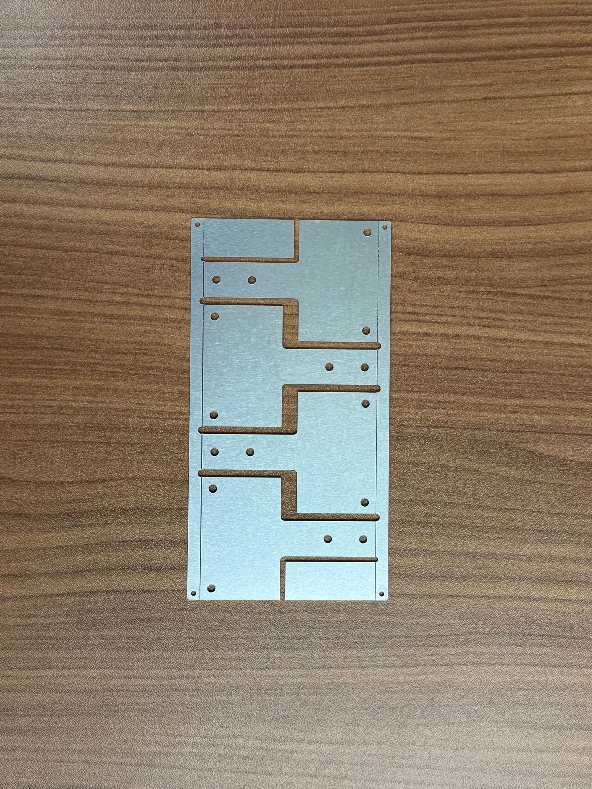 A silver-colored metal stencil with repeating geometric cutouts featuring rectangular shapes and small holes.