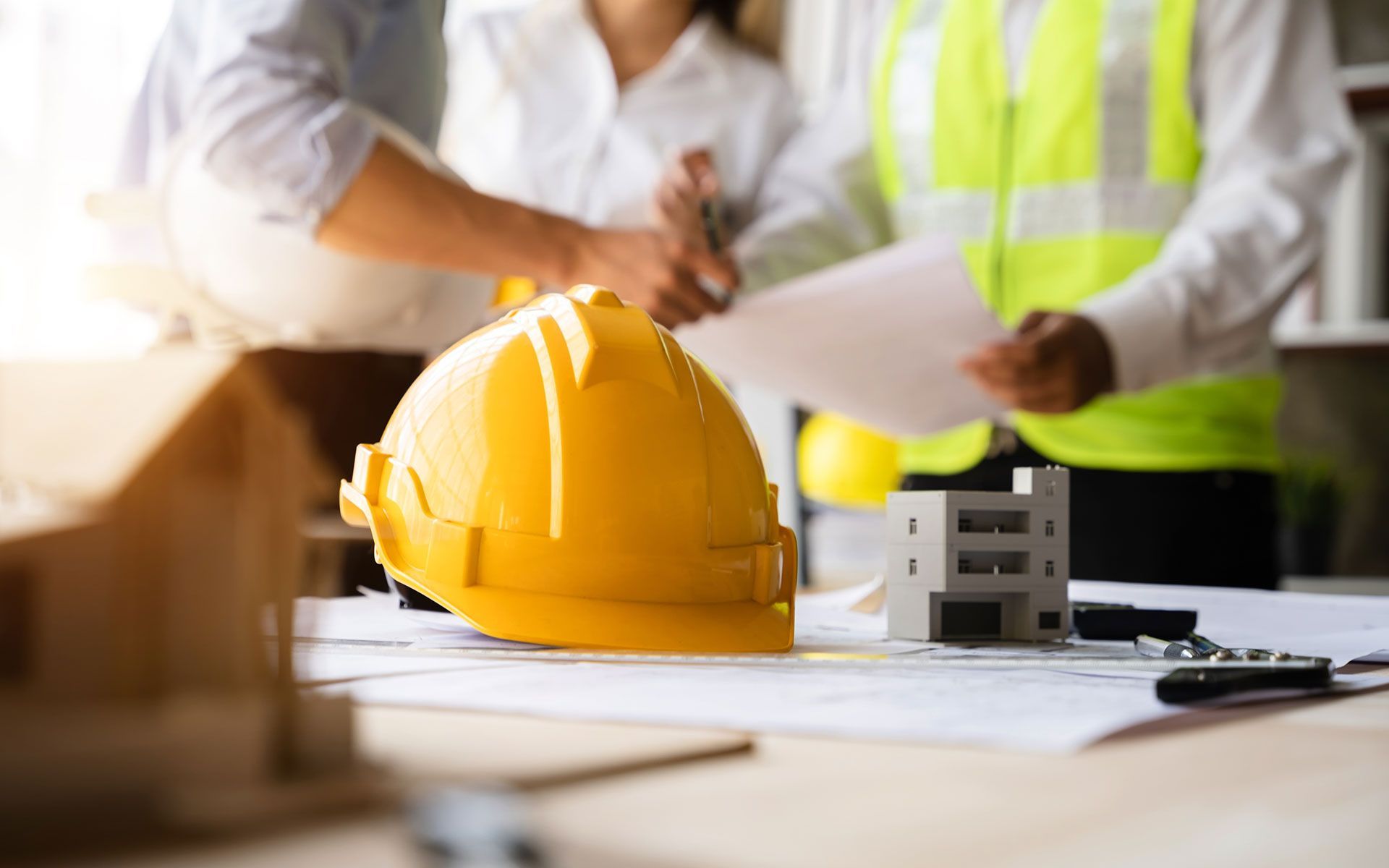 Yellow hardhat on a table with blueprints, miniature house models, and three people in the background.