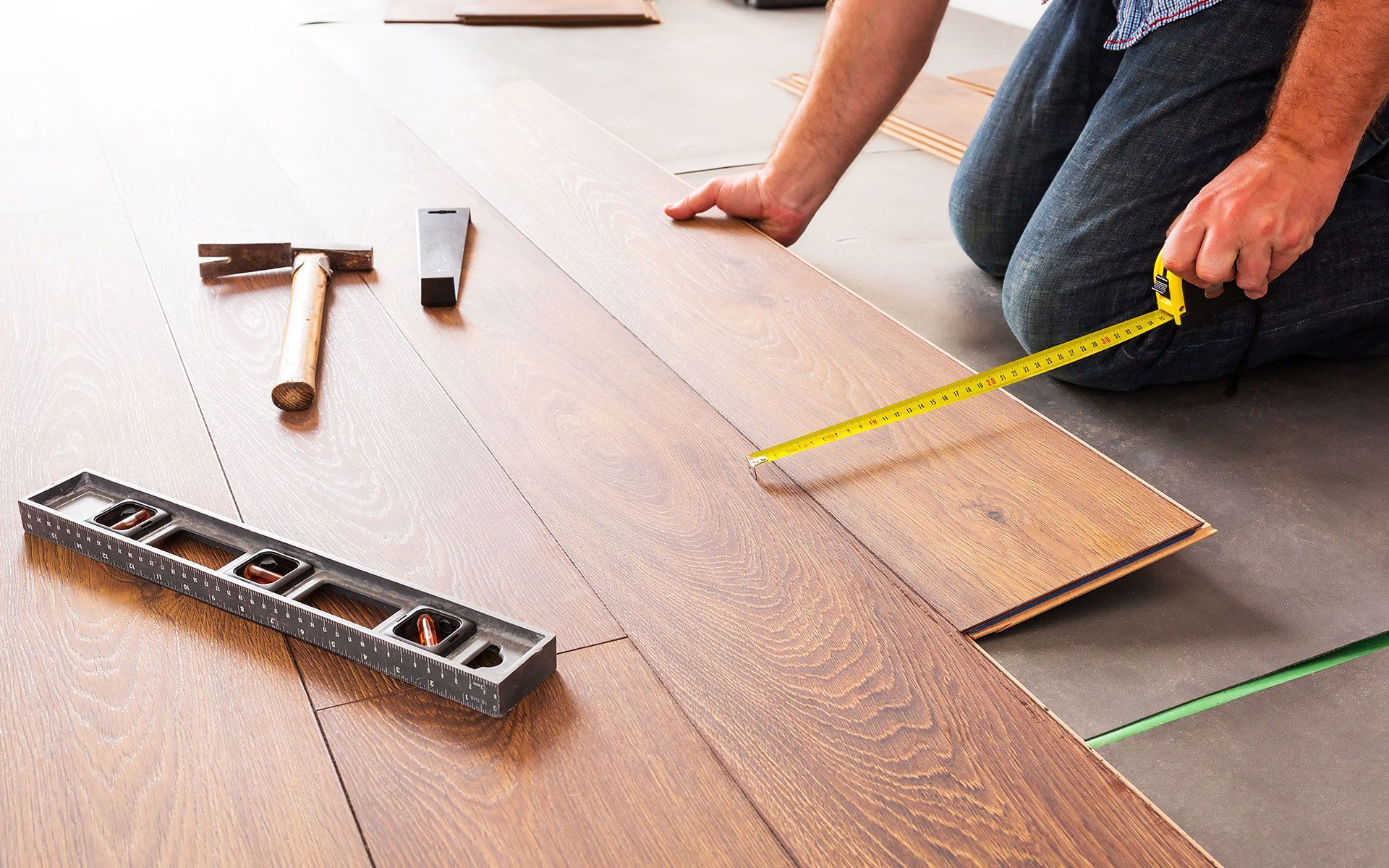Person kneeling, installing wood flooring, using measuring tape. Tools, including a hammer and level, are nearby.