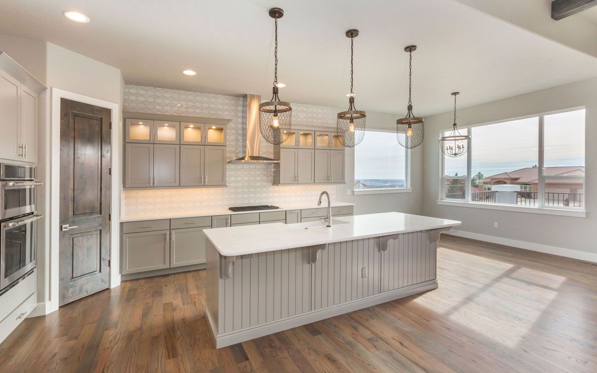Modern kitchen with island, wooden floors, gray cabinets, pendant lights, and large windows.