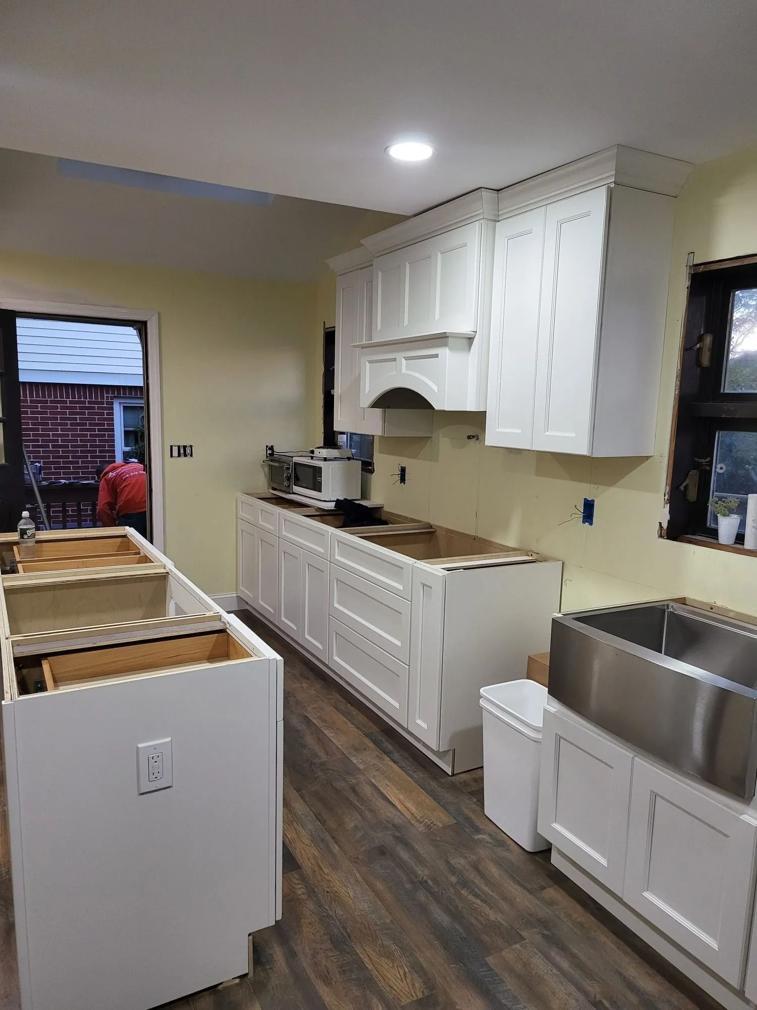 White kitchen cabinets and island under construction. Dark wood floor, light yellow walls, stainless steel sink.