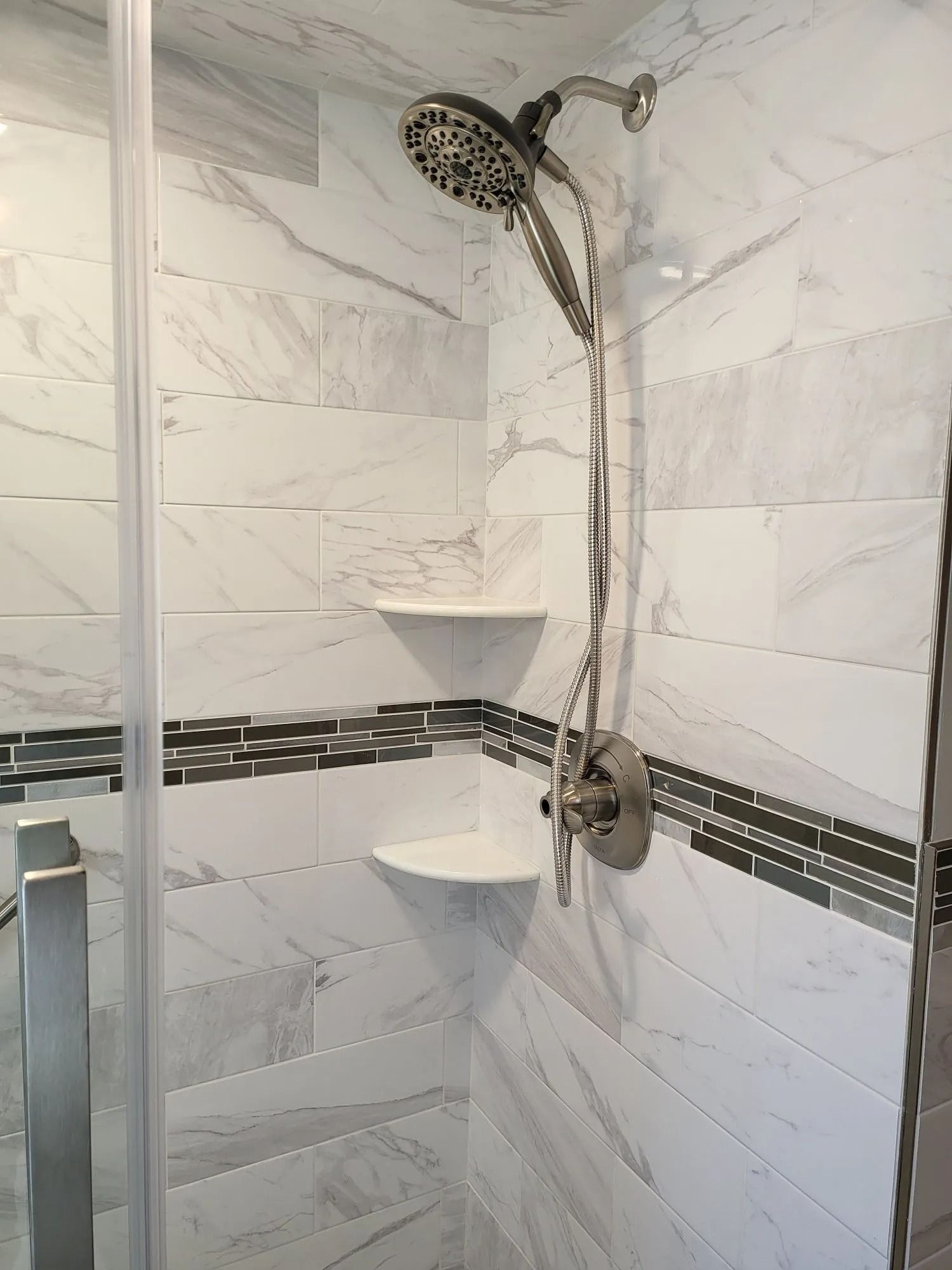 Shower with white marble-look tiles, glass door, two shelves, and dual shower heads.