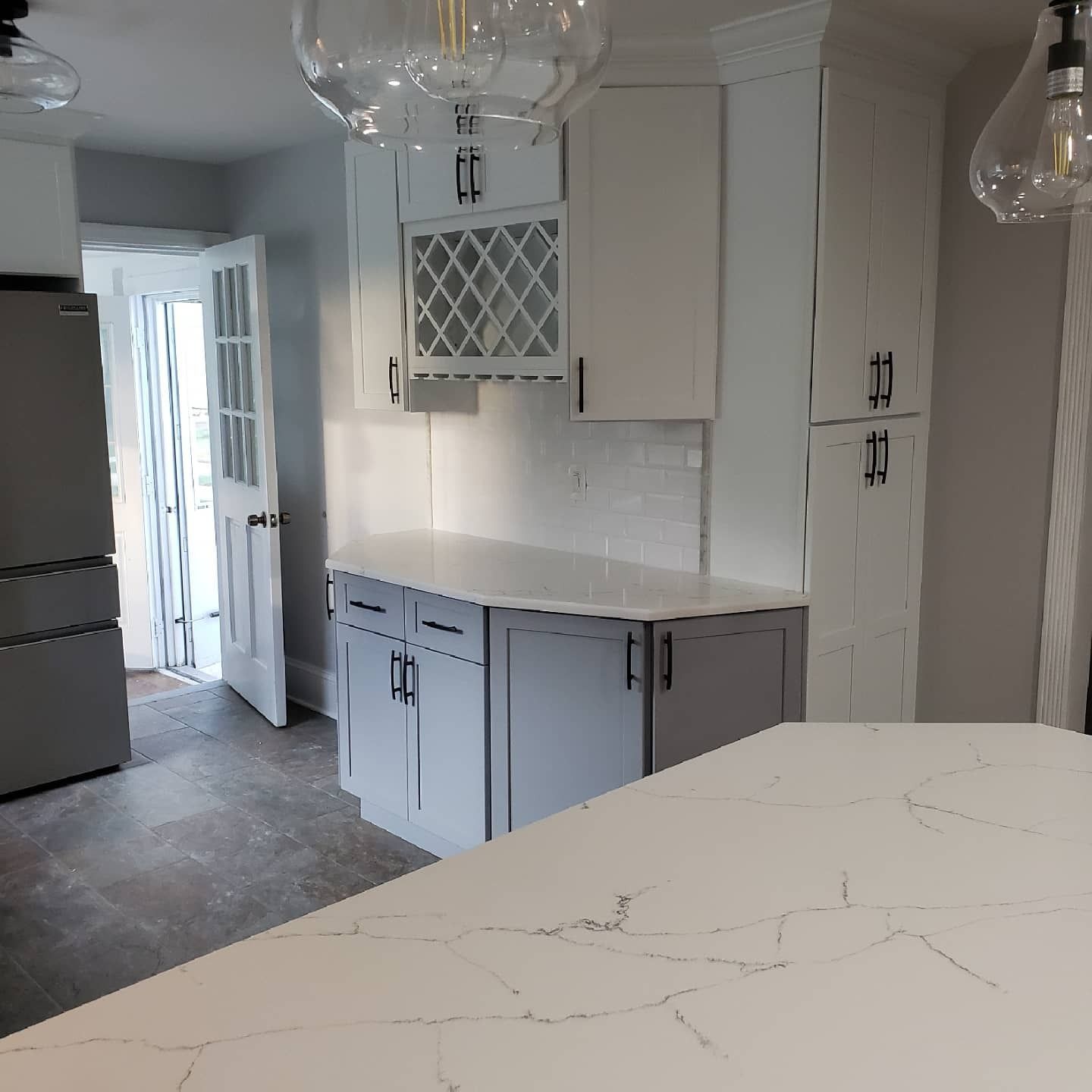 Modern kitchen with white and gray cabinets, wine rack, and white countertops. Open door on the left.