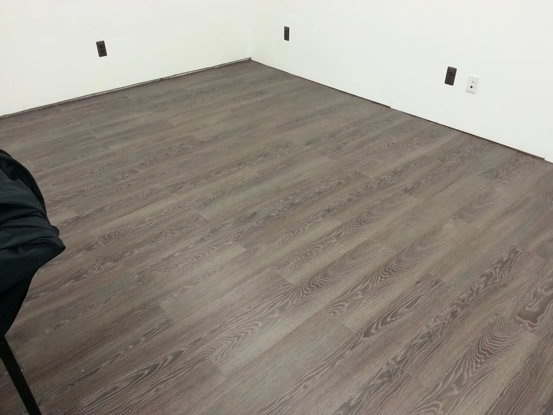 Room with gray wood-look flooring and white walls; empty corner; outlets visible.