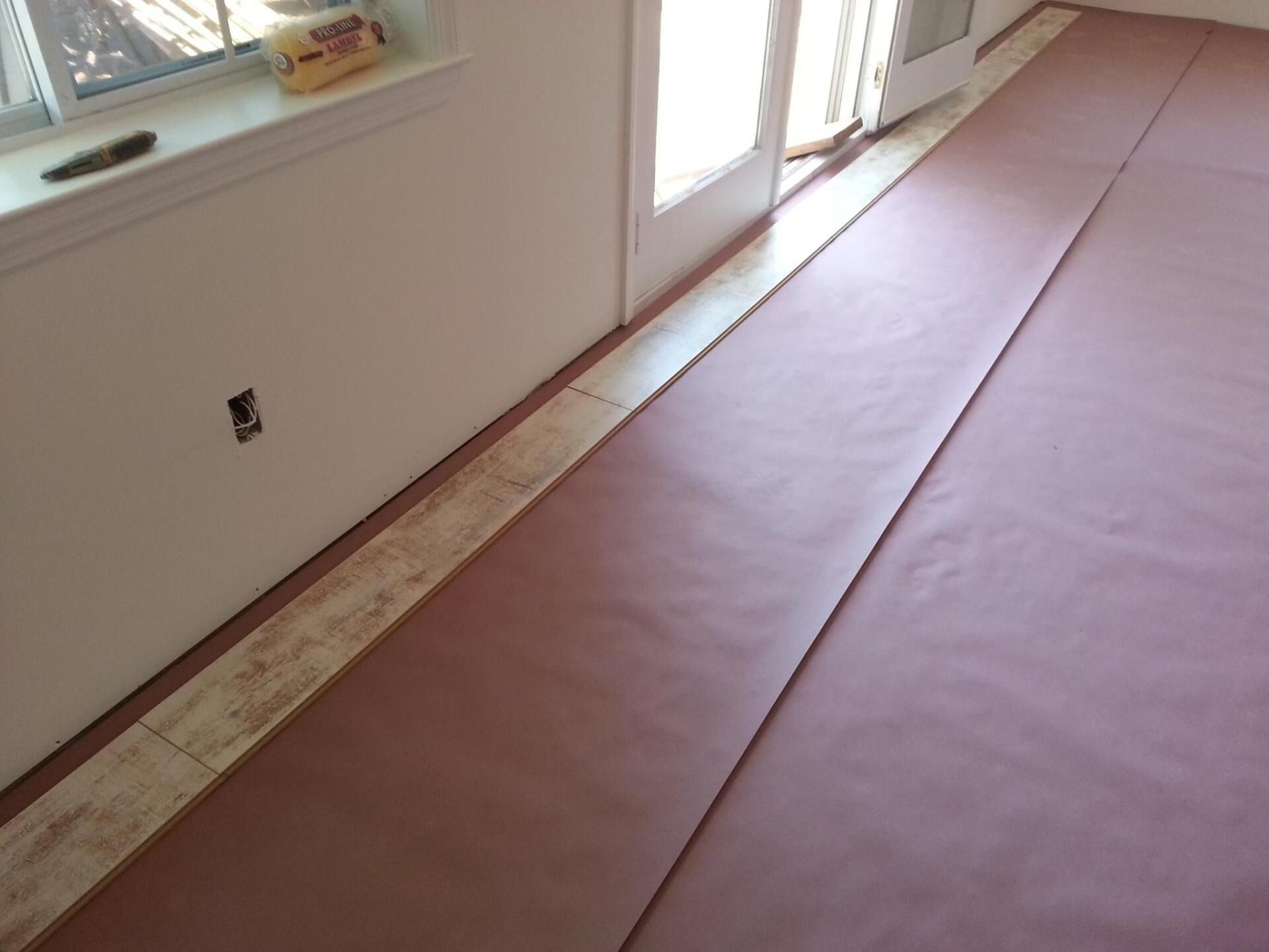 Floor installation with sheets of brown underlayment laid out. White walls, window, door.