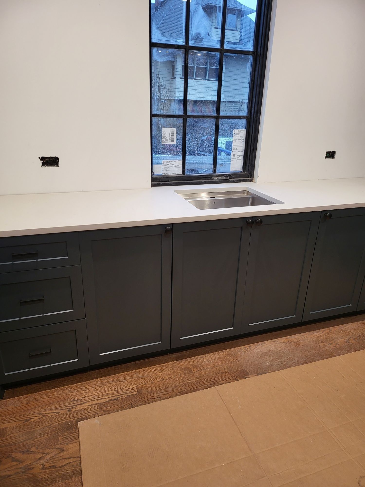 Dark gray kitchen cabinets beneath a white countertop and a window with a stainless steel sink.
