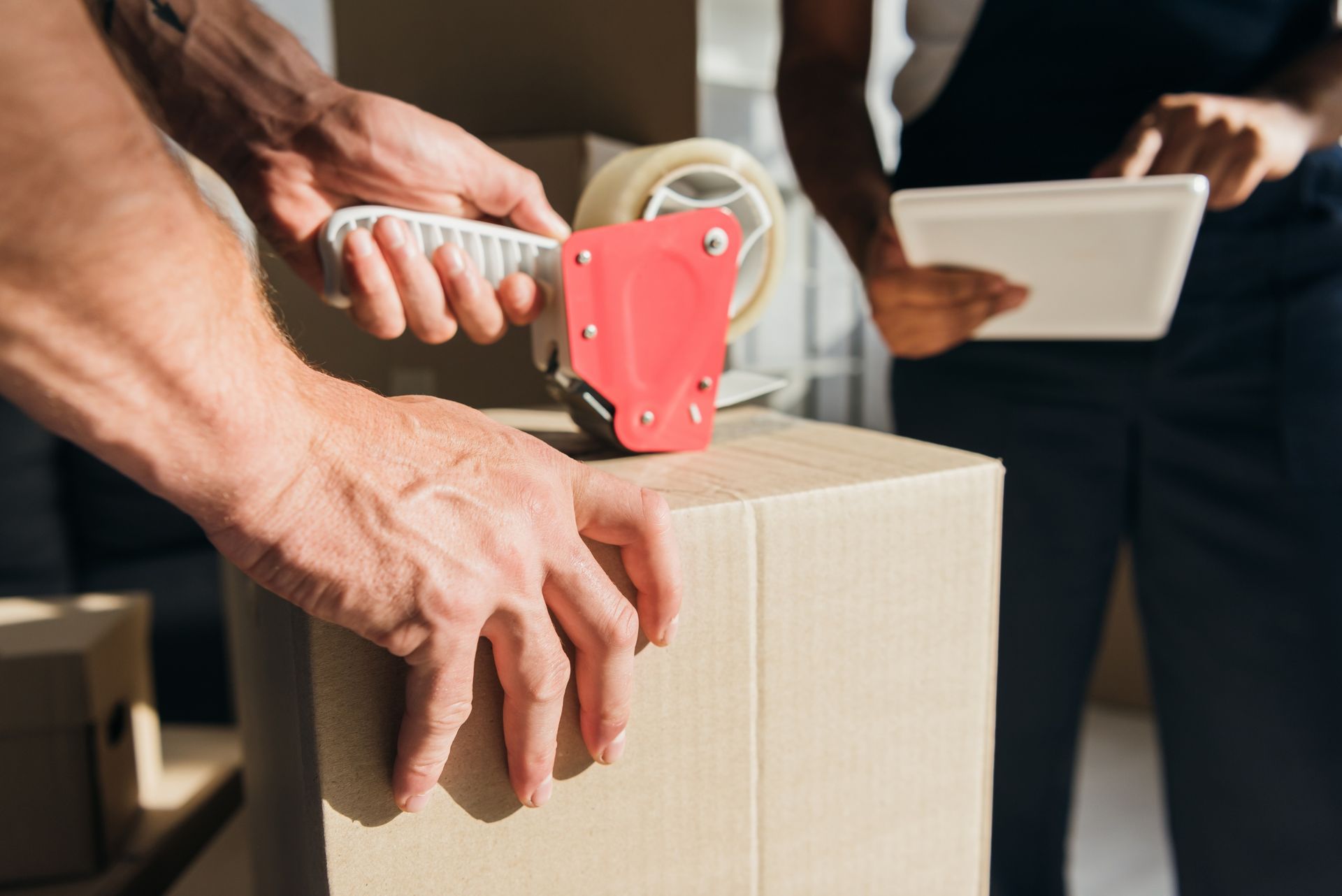 Hands sealing a cardboard box with red tape while a person holds a clipboard nearby.