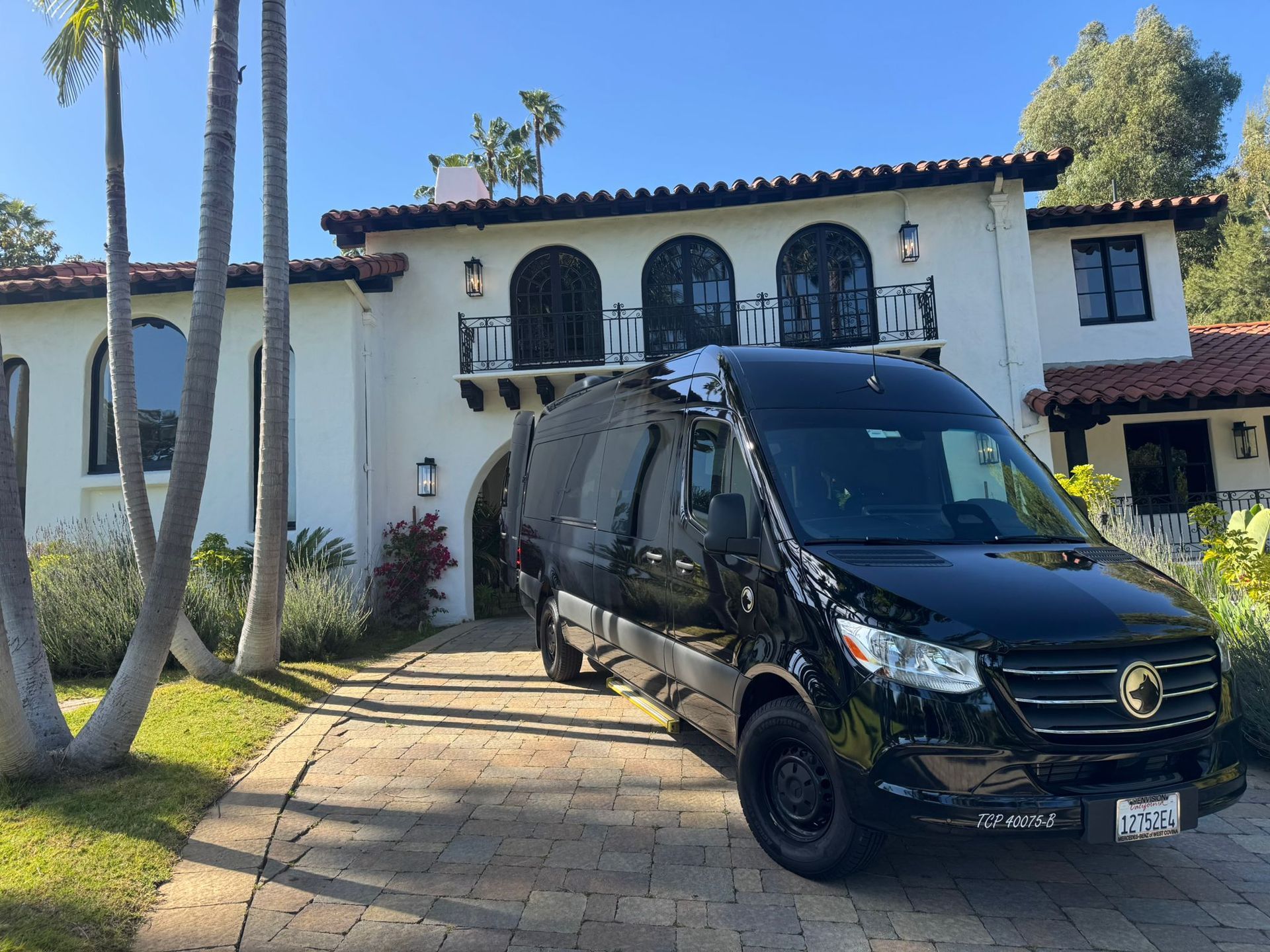 Black van parked in front of a white Spanish-style house with palm trees and a sunny lawn.