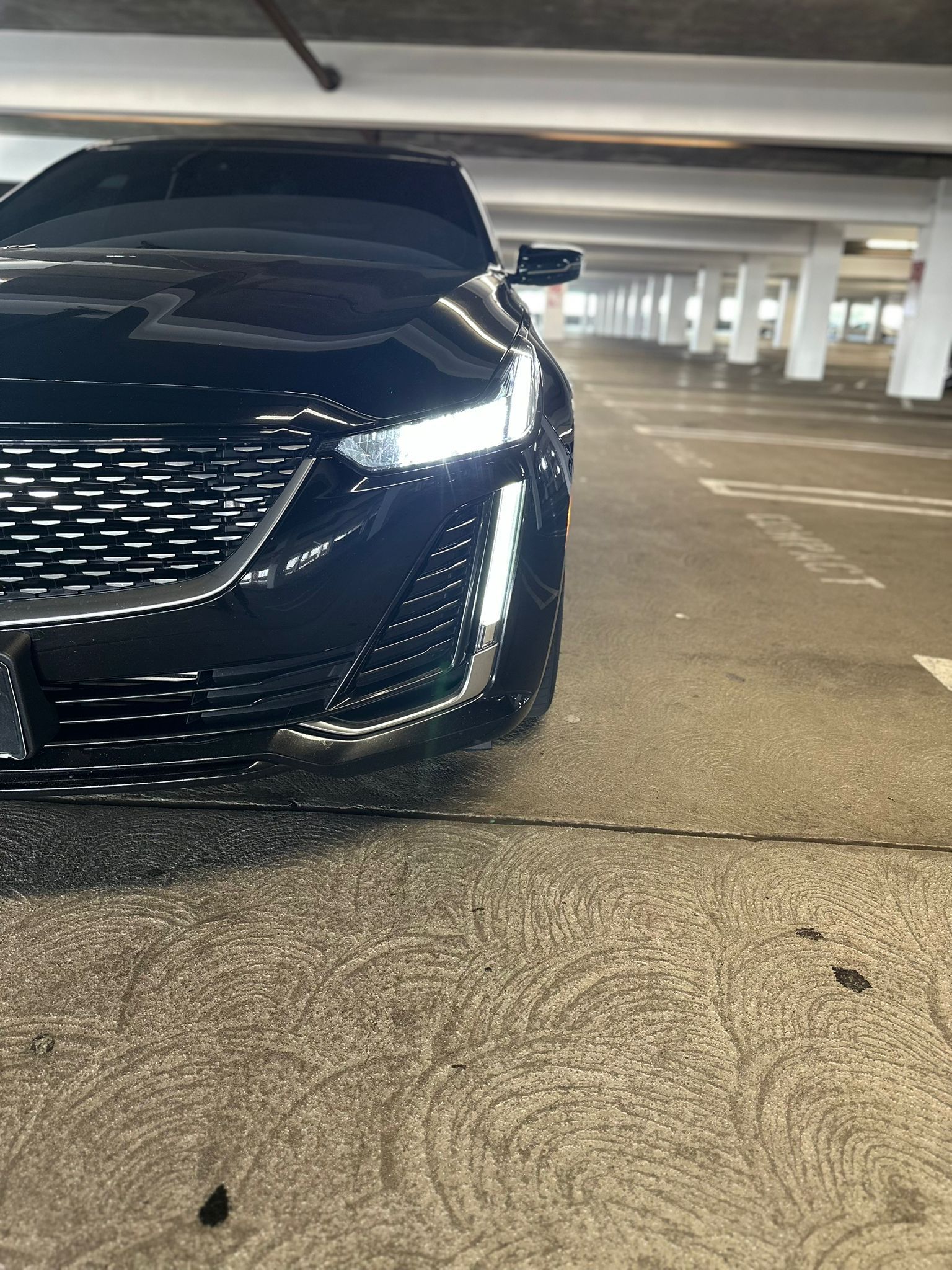 Black sedan parked in a concrete parking garage, front-left view with headlights on