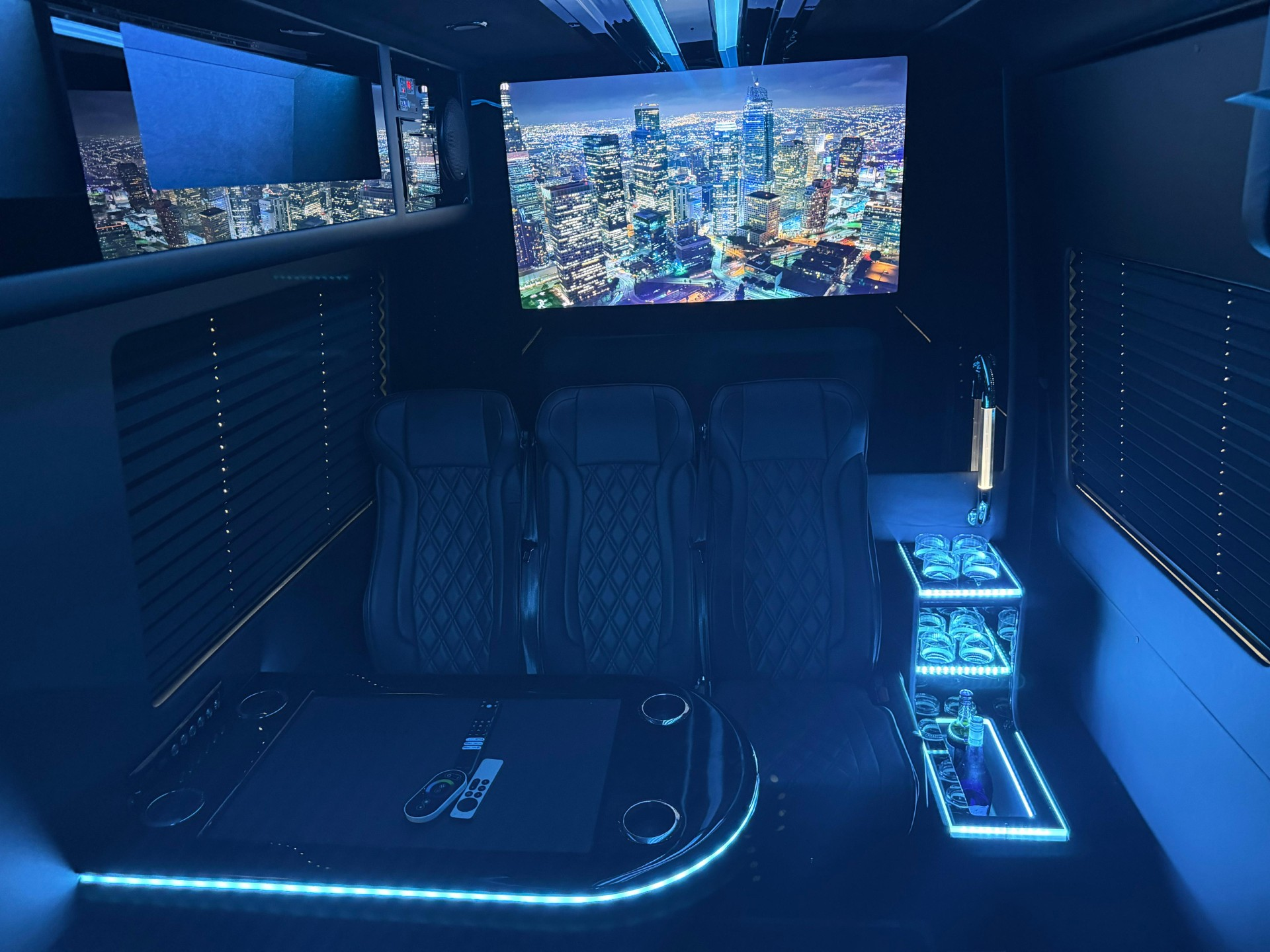 Blue-lit luxury van interior with rear seats, panoramic city view screens, and a small illuminated table