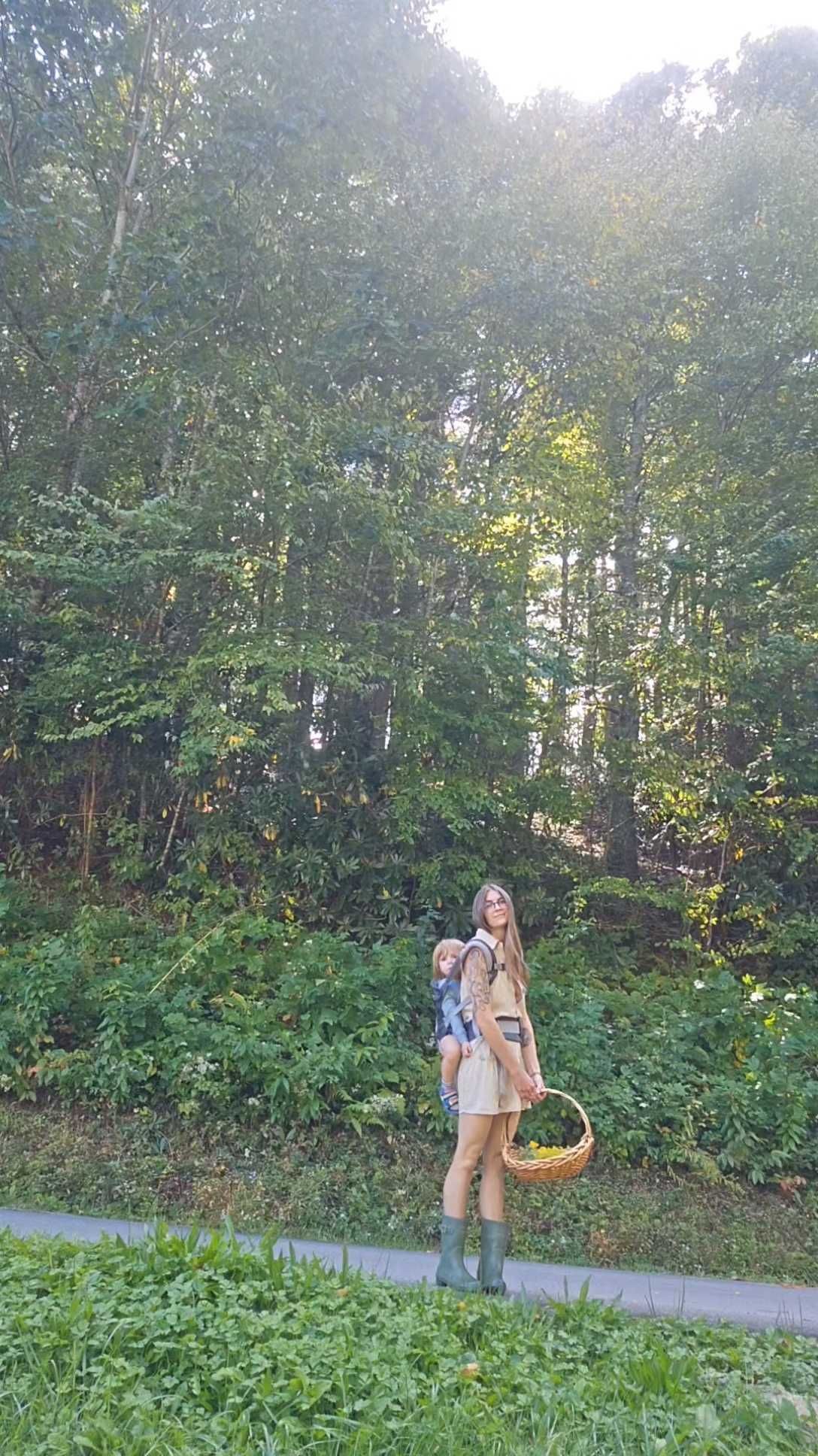 Person standing on a path in front of dense trees, holding a woven basket in a sunlit forest.
