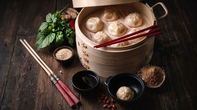 Steamed buns in a bamboo basket with chopsticks, soy sauce, and herbs on a dark wooden table