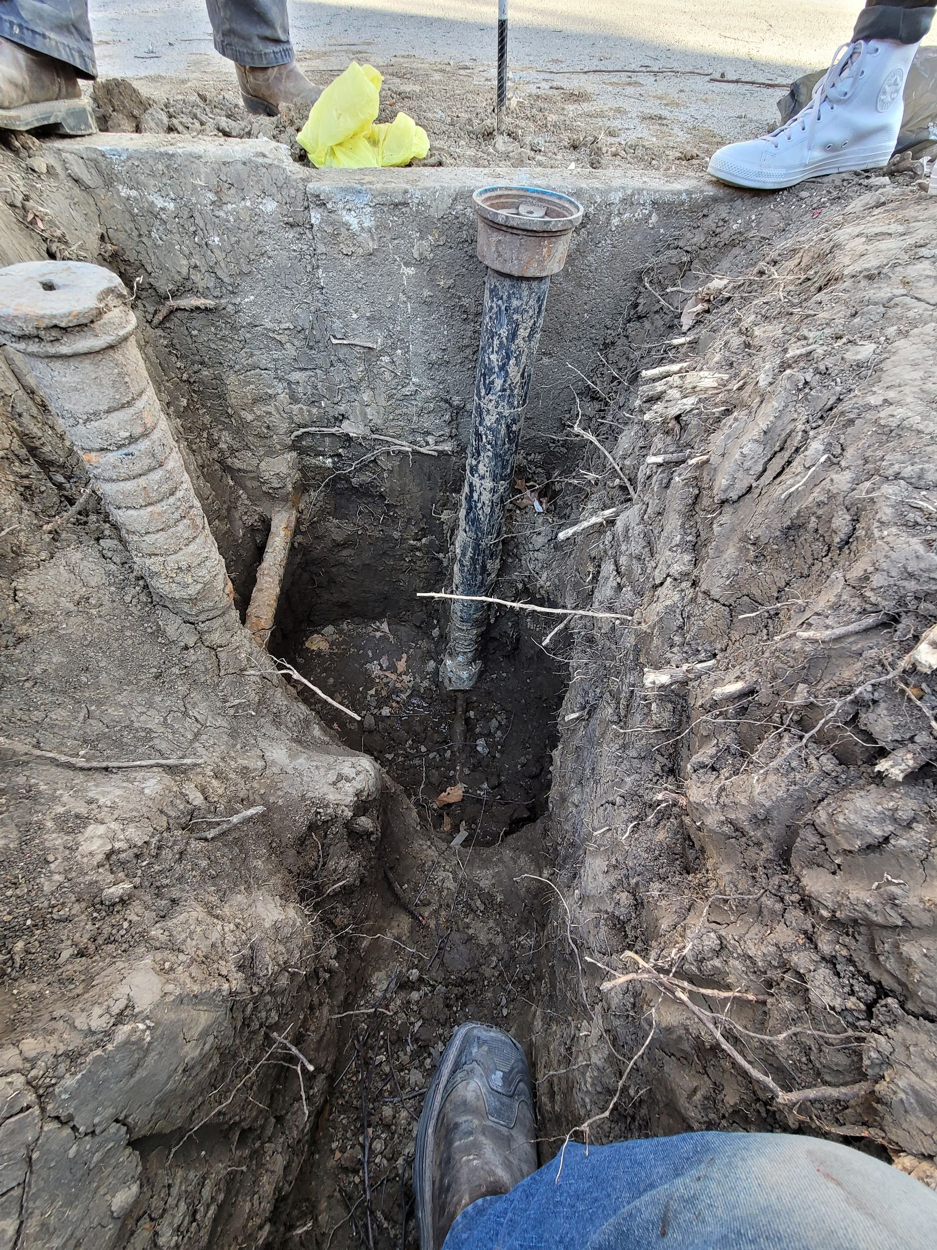 A water utility worker stands in a dirt trench, inspecting an exposed vertical pipe leaking water near a concrete curb.