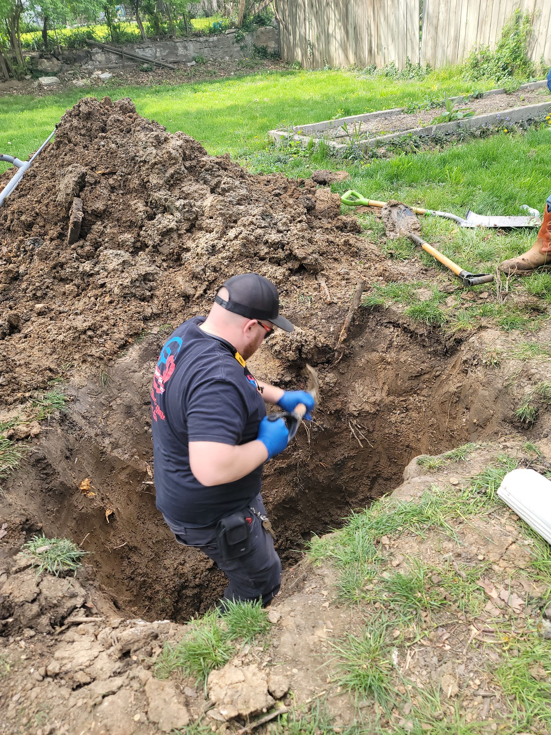 A person in a black shirt and cap uses a small shovel to dig in a rectangular trench outdoors next to a pile of dirt.