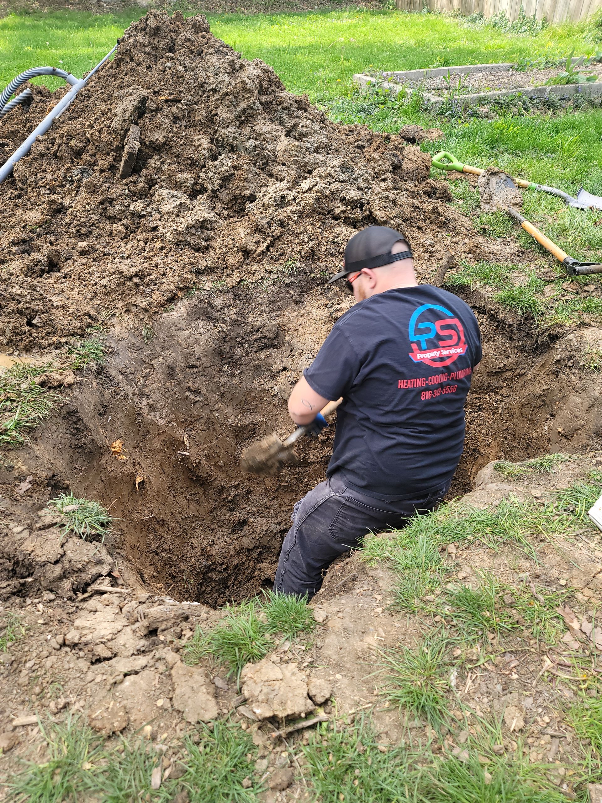 A person wearing a dark t-shirt and baseball cap shovels dirt out of a deep rectangular hole in a grassy yard.