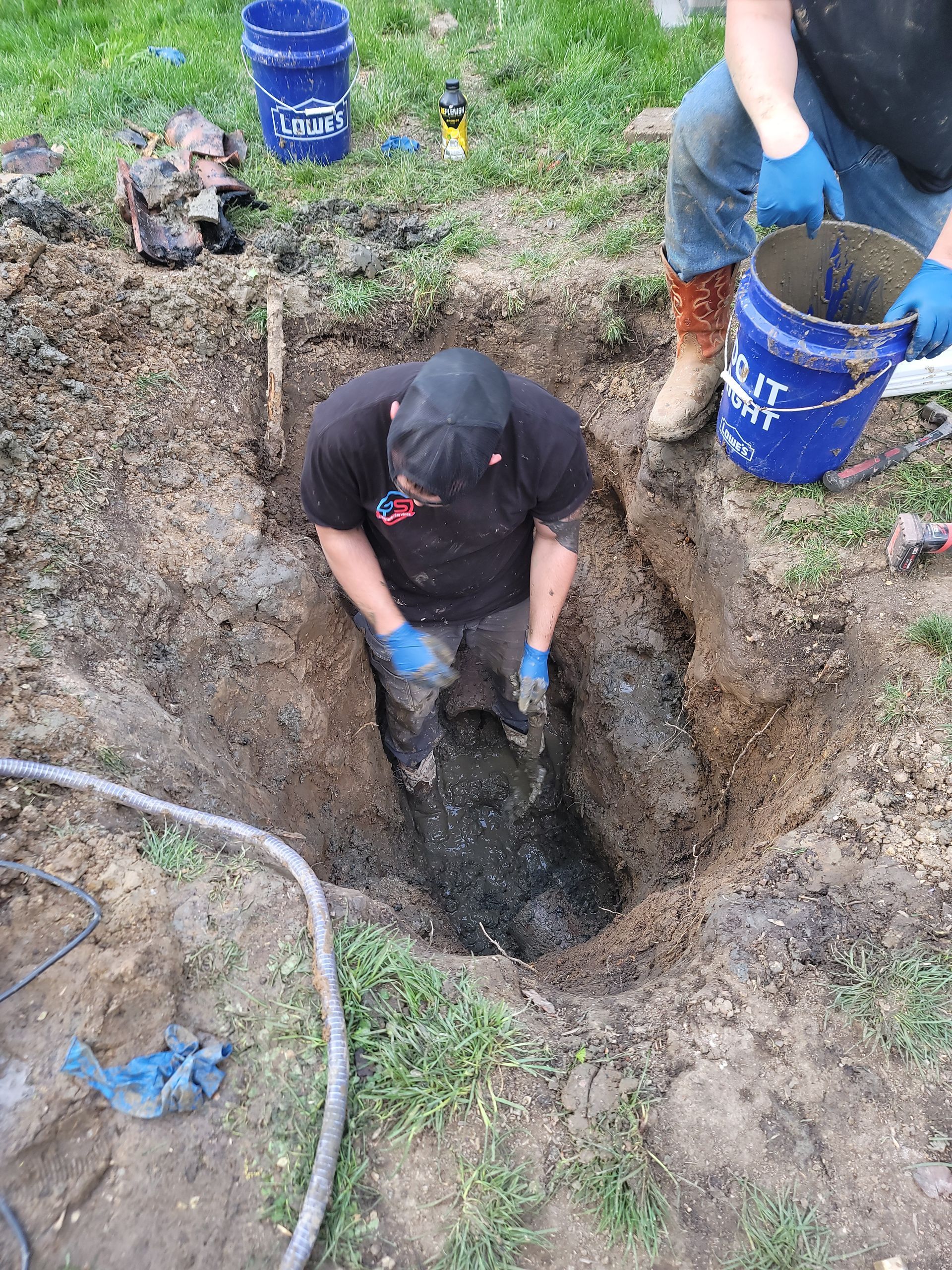 A person in a muddy pit works on underground pipes, with a second person standing nearby holding a blue bucket.