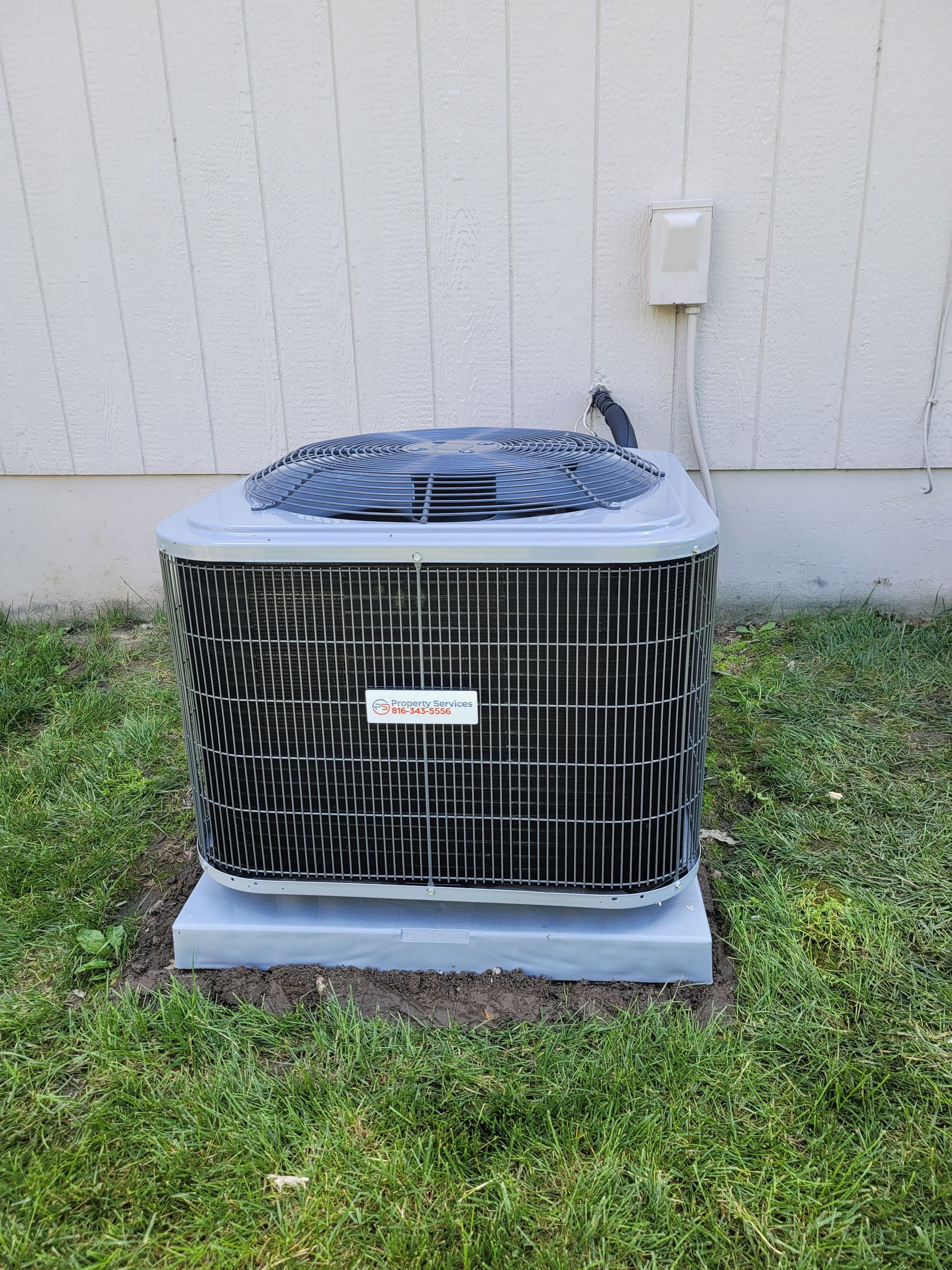 A grey HVAC condenser unit sits on a concrete pad against a beige house wall with green grass in the foreground.