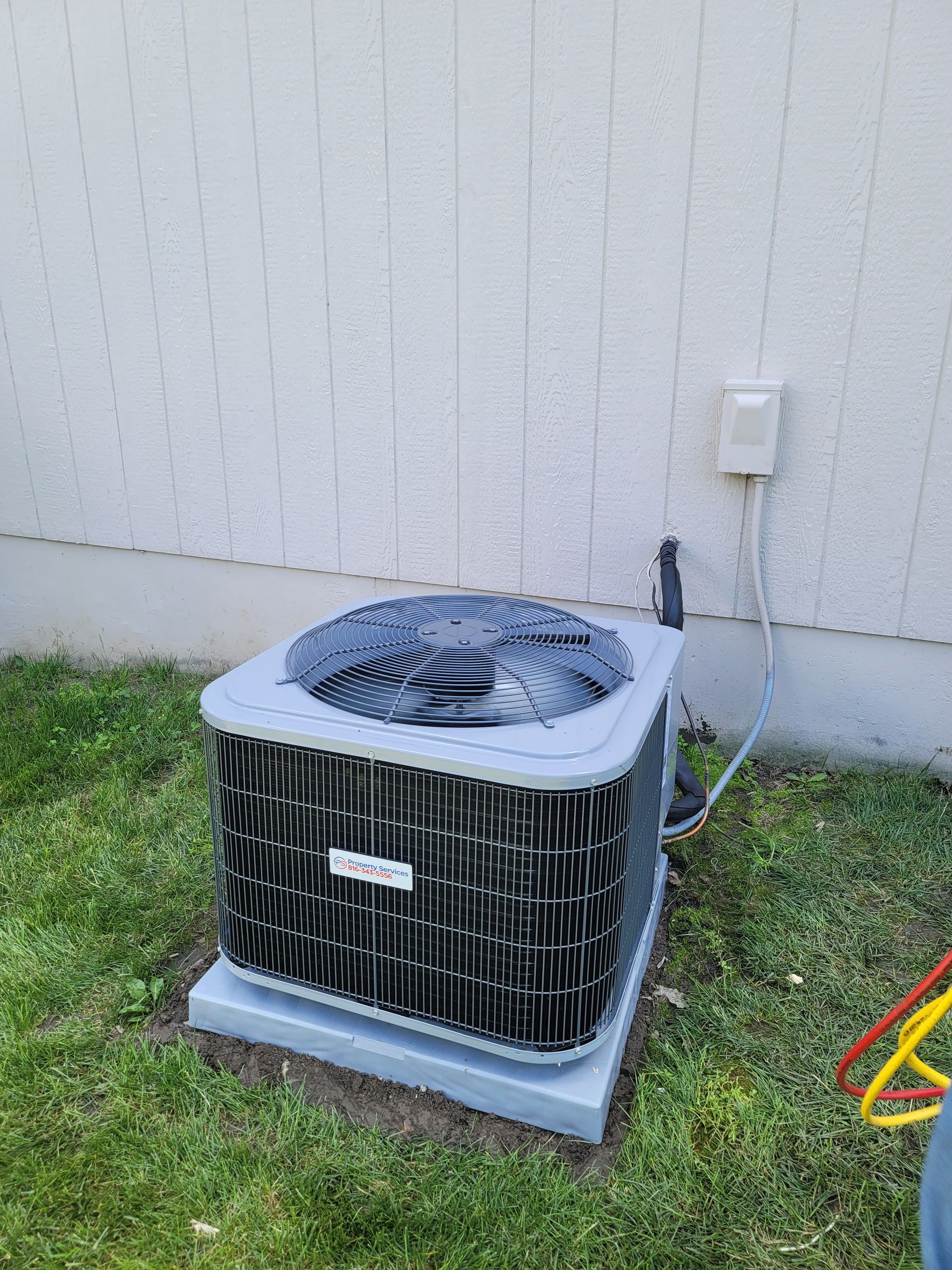 A grey, square HVAC condenser unit sits on a concrete pad in a grassy yard against a white vertical-siding wall.