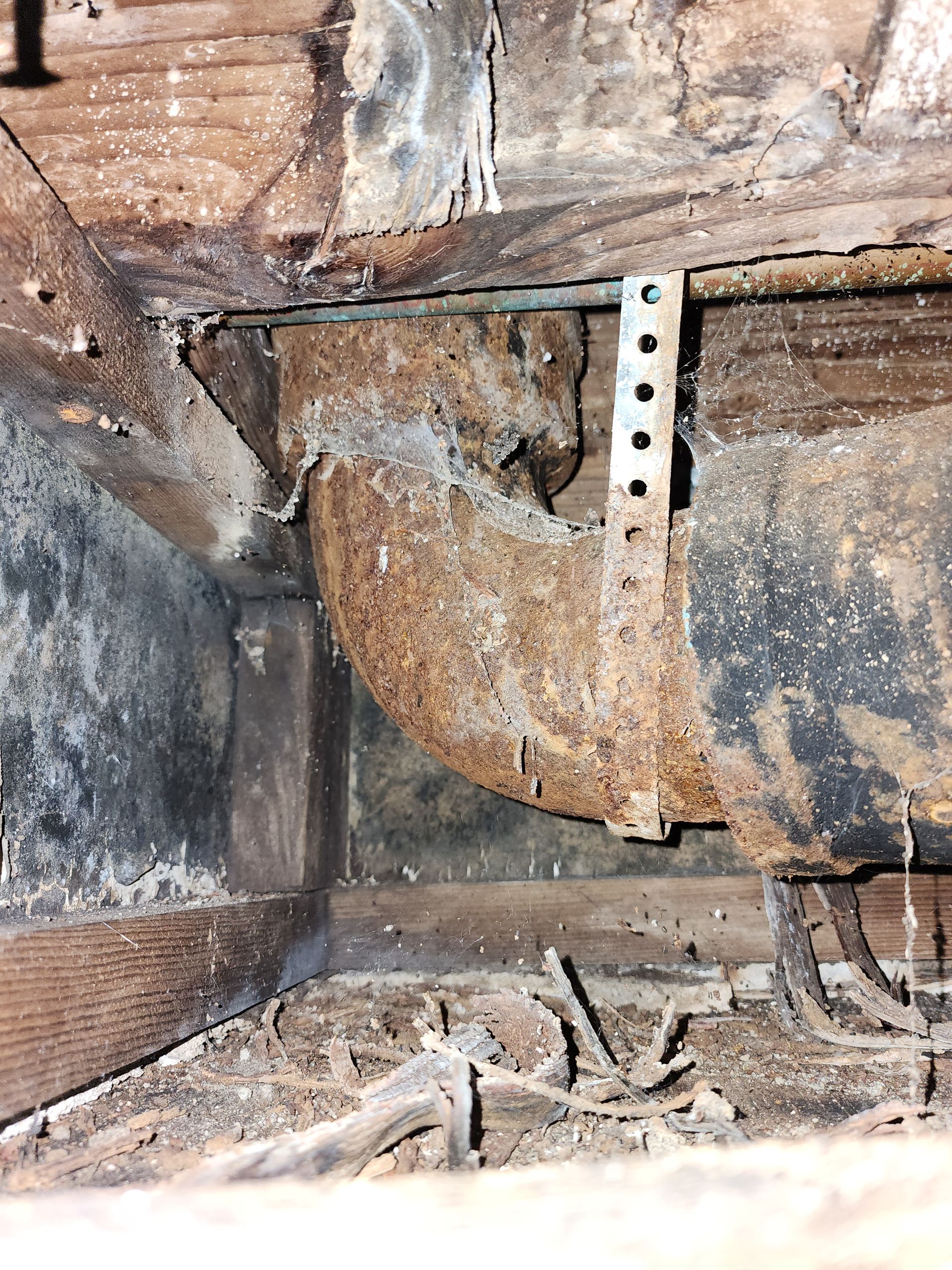 A rusted metal drain pipe secured by a perforated steel strap beneath a wooden subfloor covered in dust and debris.