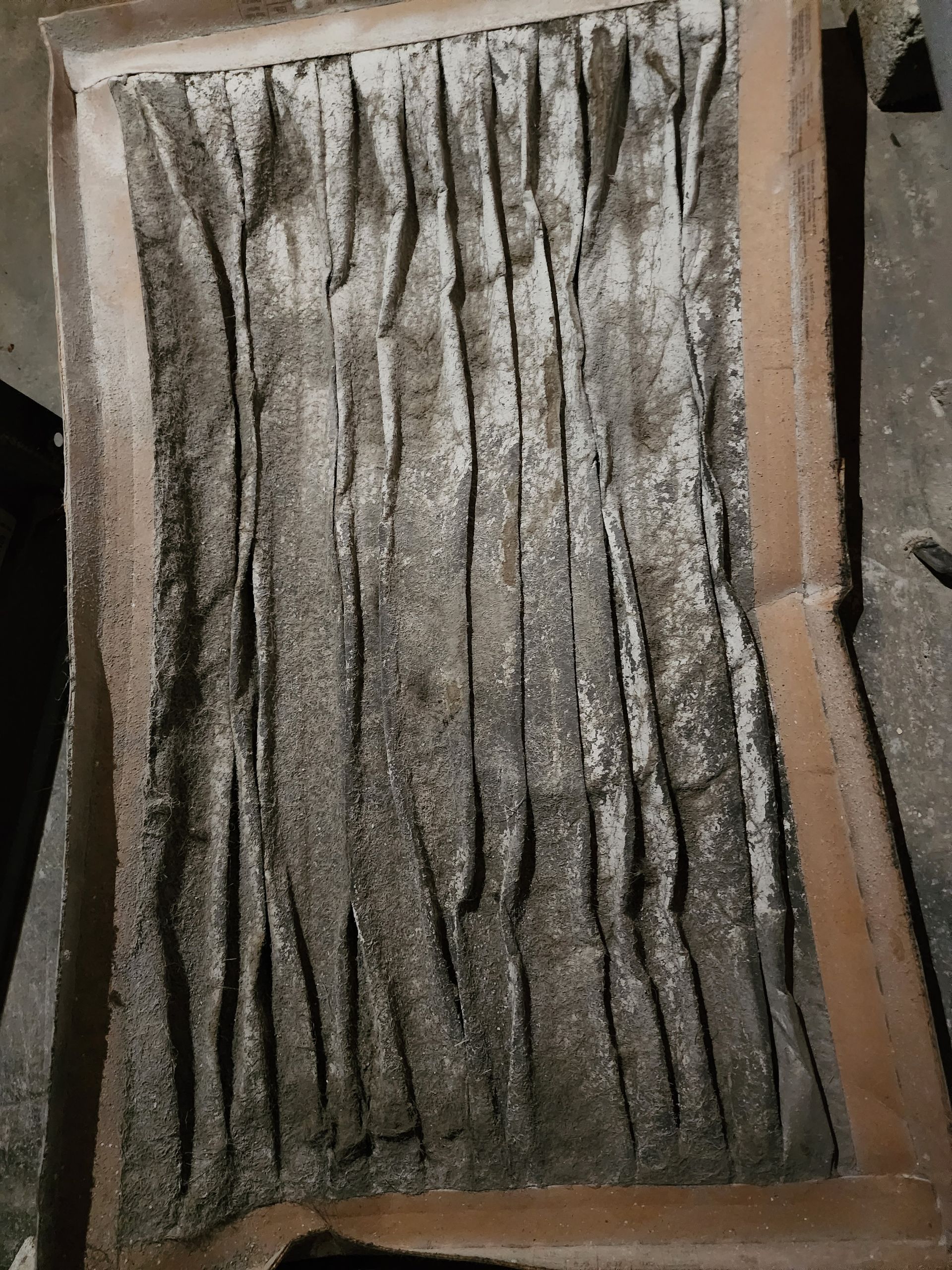 A rectangular HVAC air filter covered in a thick layer of grey dust and debris, showing heavy use and blockage.