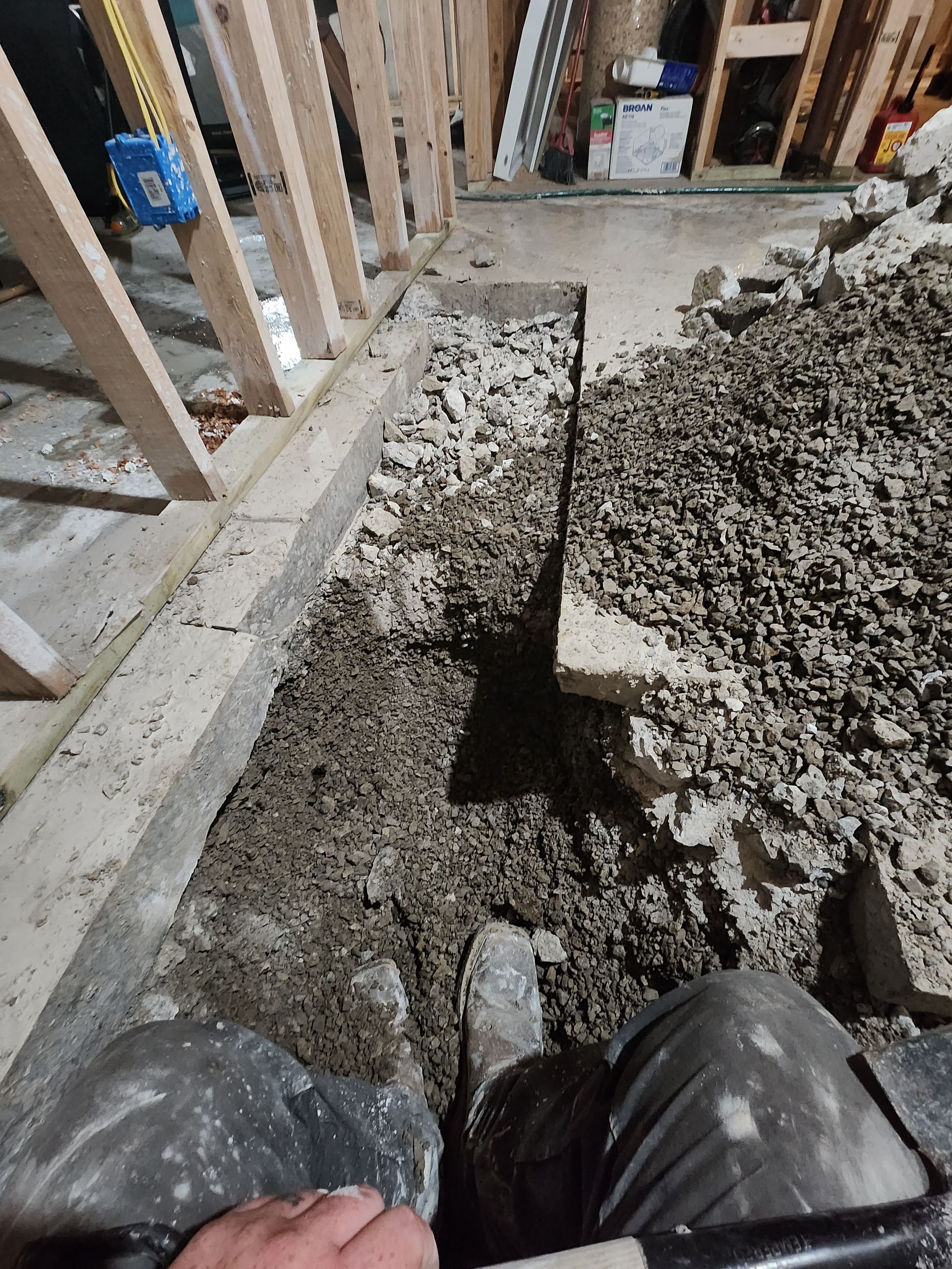 A view from above showing a trench dug through a concrete floor next to wooden wall studs in a construction setting.