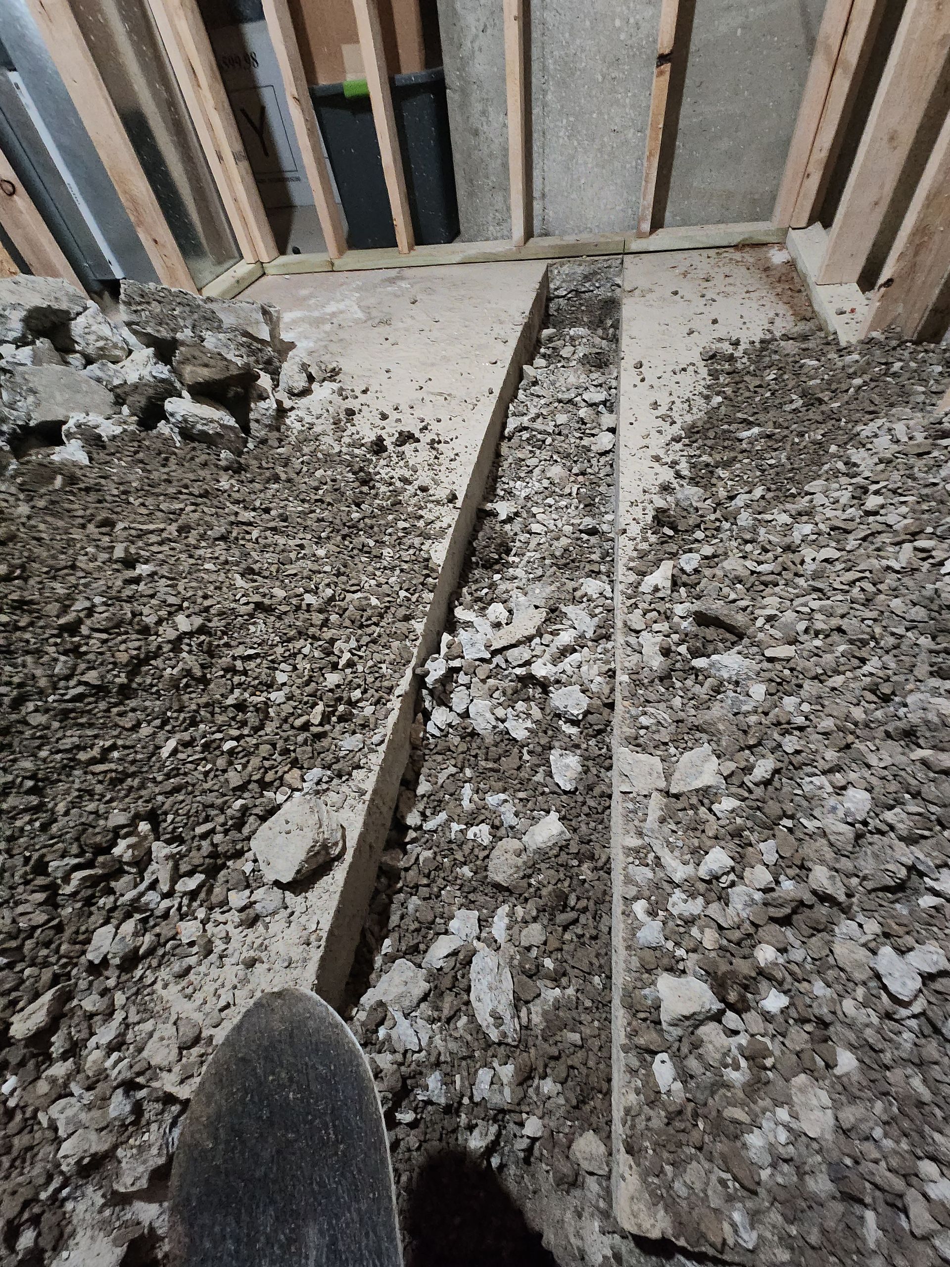 A narrow trench cut into a concrete basement floor, surrounded by debris and wooden wall framing.