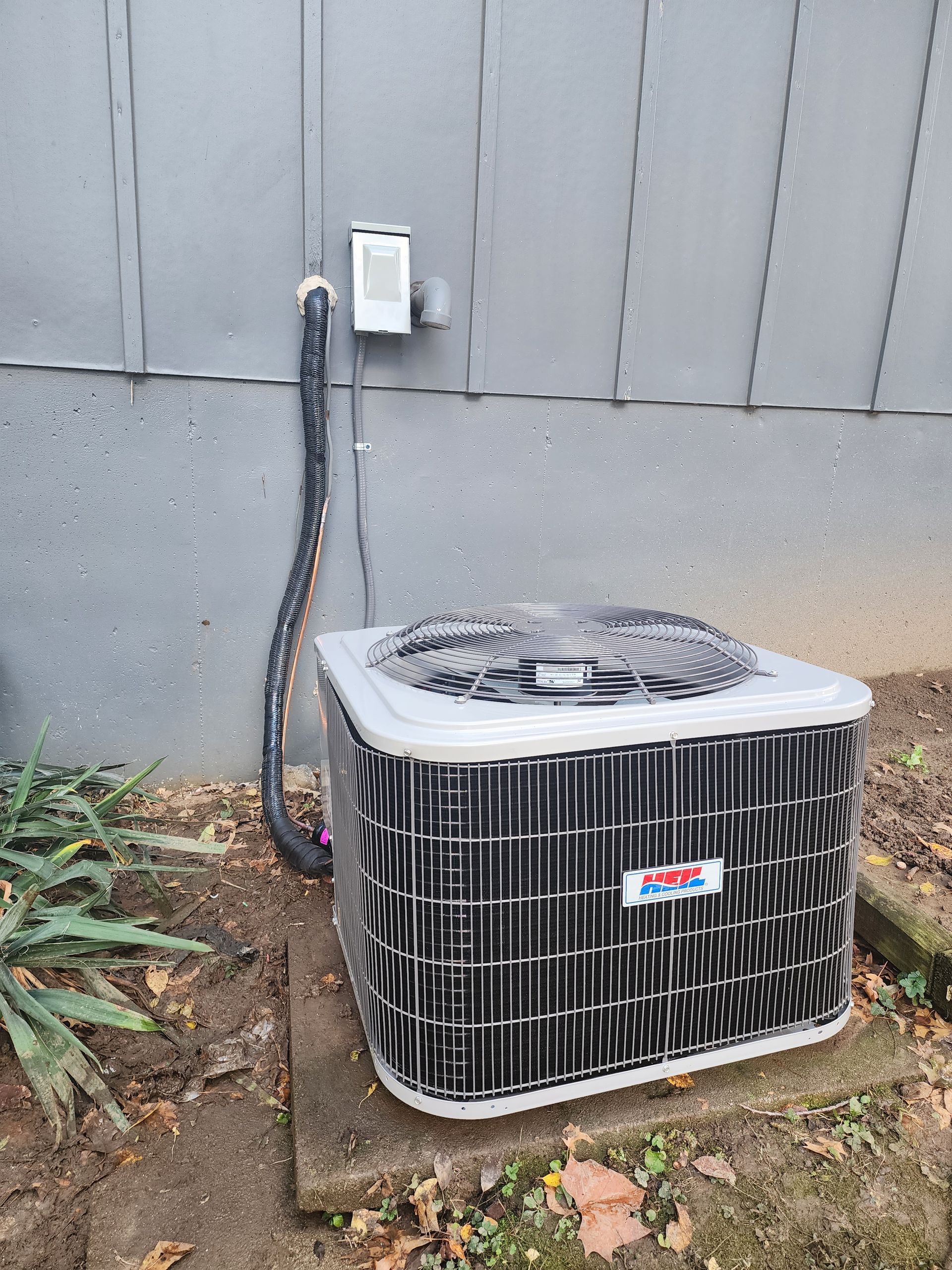 An outdoor air conditioning condenser unit sits on a concrete pad against a gray exterior wall.