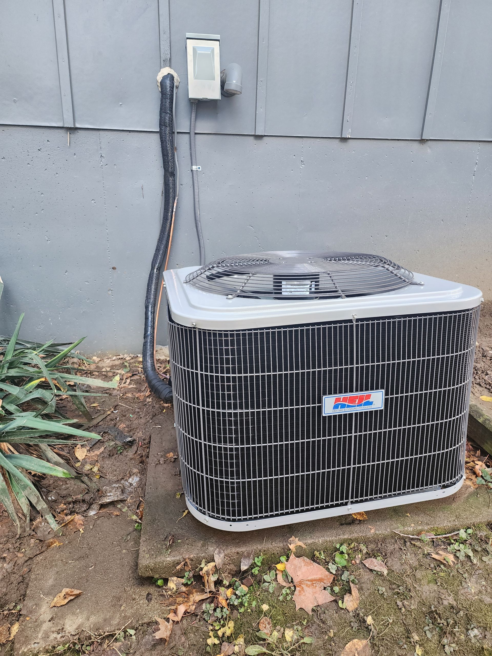 An outdoor air conditioning condenser unit sitting on a concrete pad against a gray wall with an electrical disconnect.