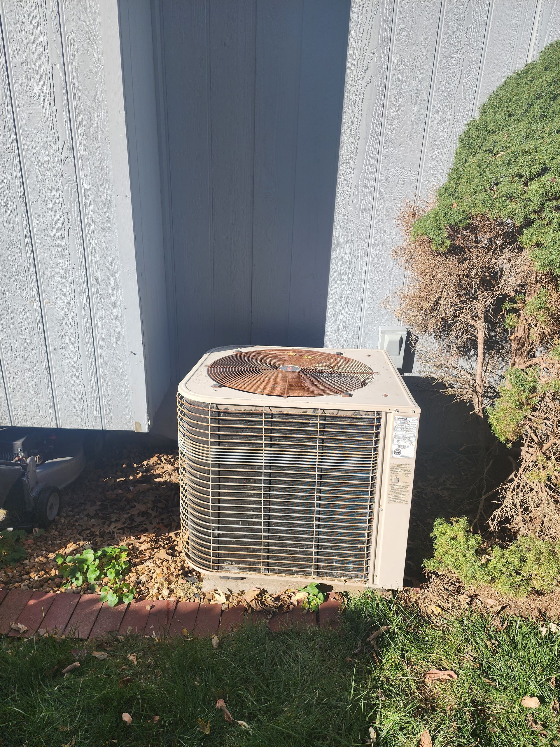 A weathered, square air conditioning condenser unit sits on wood chips next to a gray wall and a green bush.