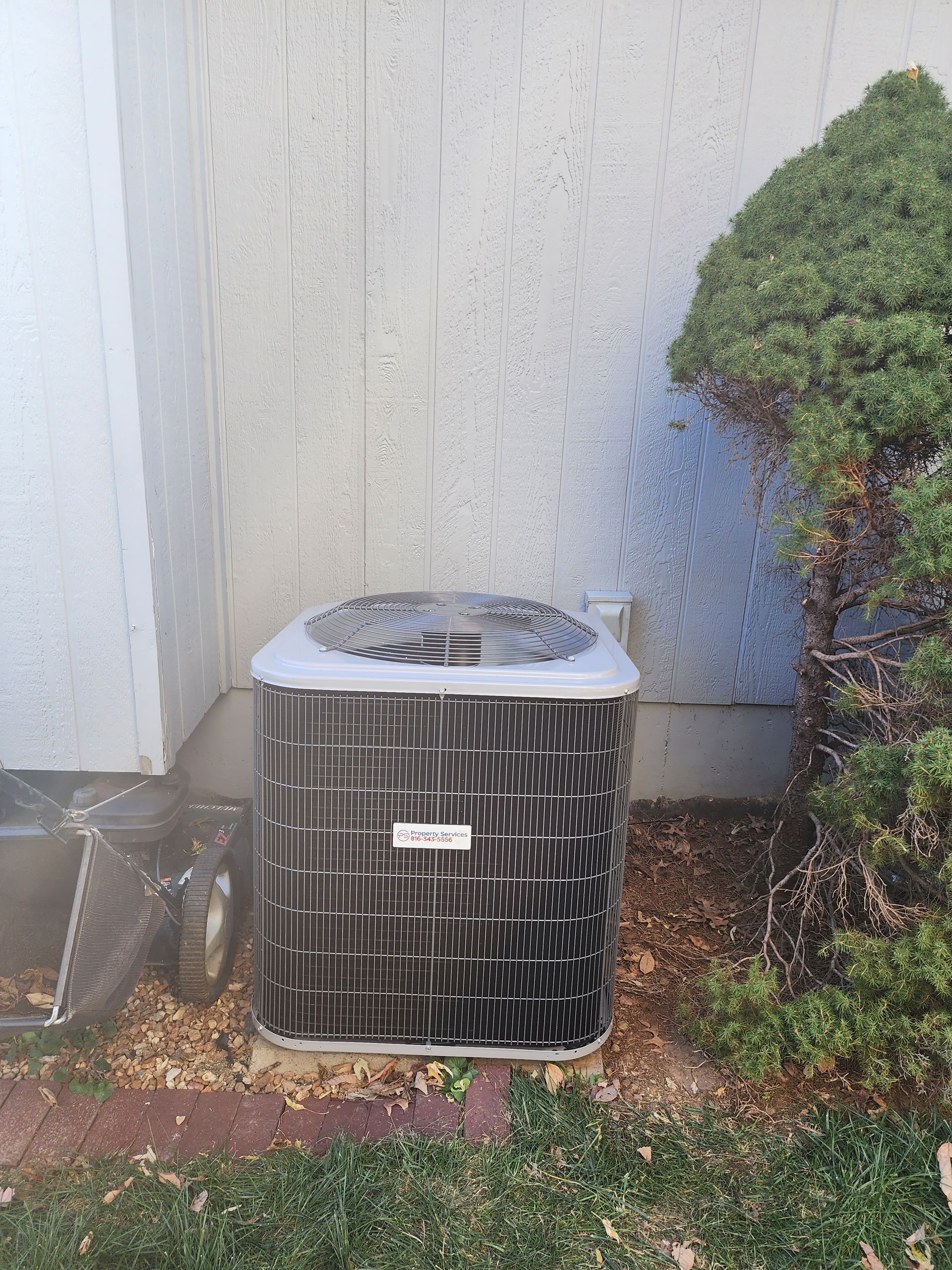 An outdoor residential air conditioning unit sitting next to a beige house wall and a green bush.